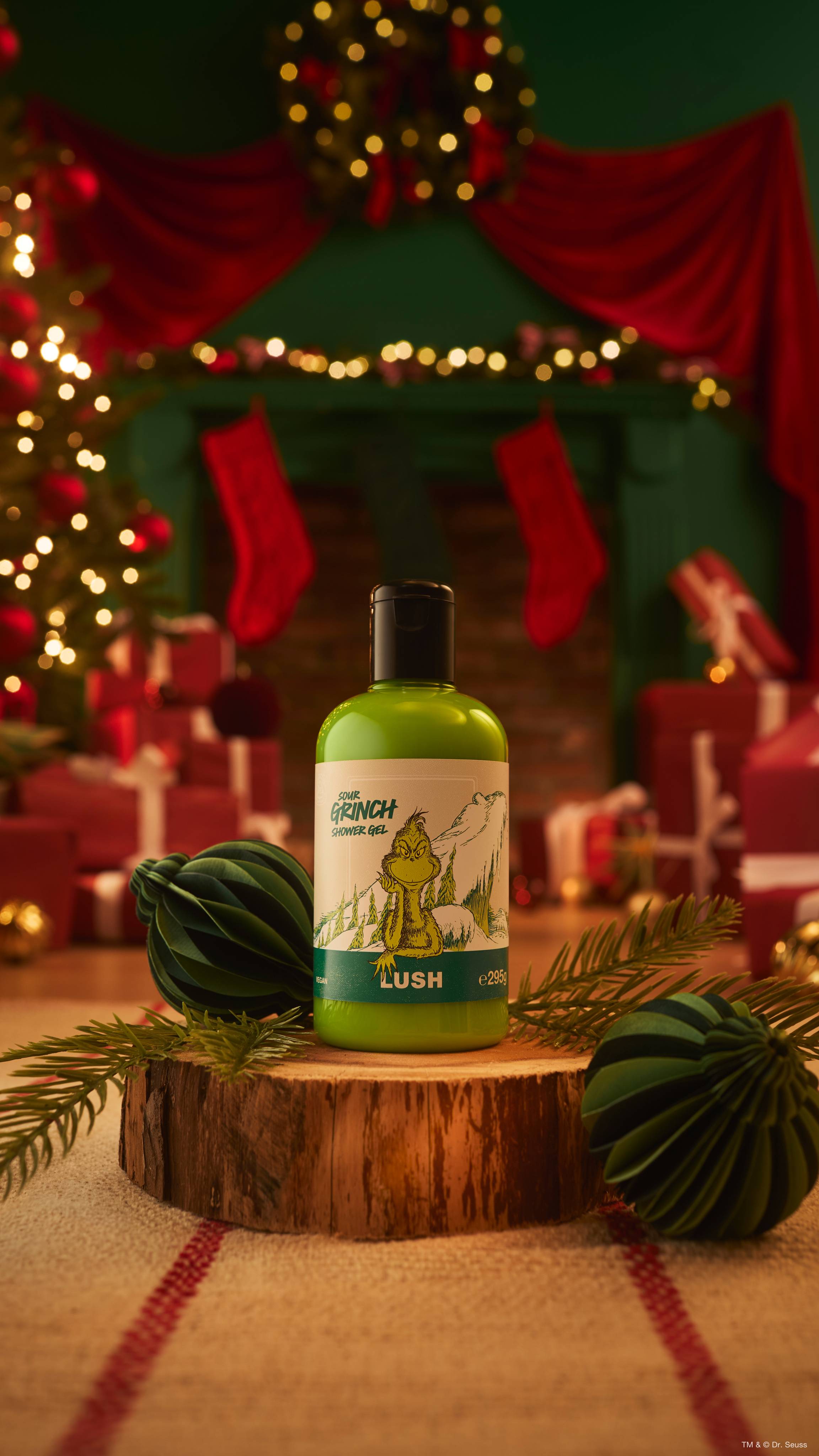 A bottle of Lush 'Sour Grinch' bright green shower gel sits on a wooden log slice flanked by green paper baubles, set against a cozy, blurred Christmas fireplace backdrop decorated with lights and red drapes.  