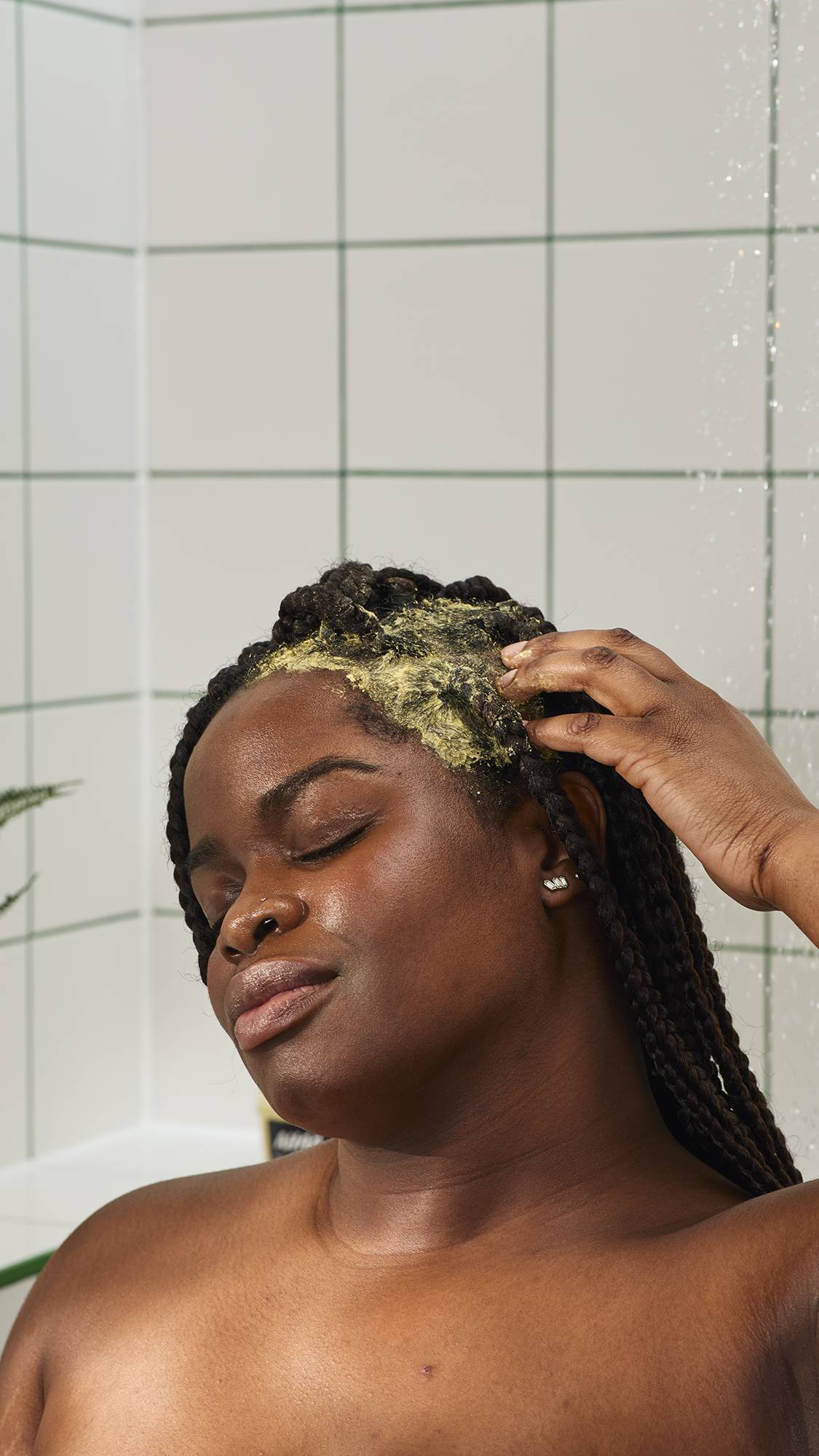 A person with long dark braids leans their head back with eyes closed, massaging a thick, mustard-yellow treatment into their scalp and hair. They are in a shower with white tiles and green grout, with water falling in the background.