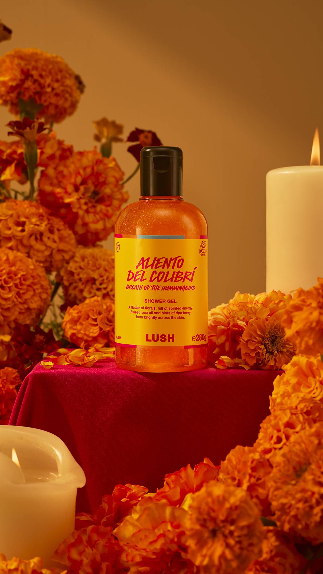 A bottle of Lush Aliento Del Colibrí shower gel sits on a red draped pedestal, surrounded by vibrant orange marigold flowers and lit candles in a warm display.