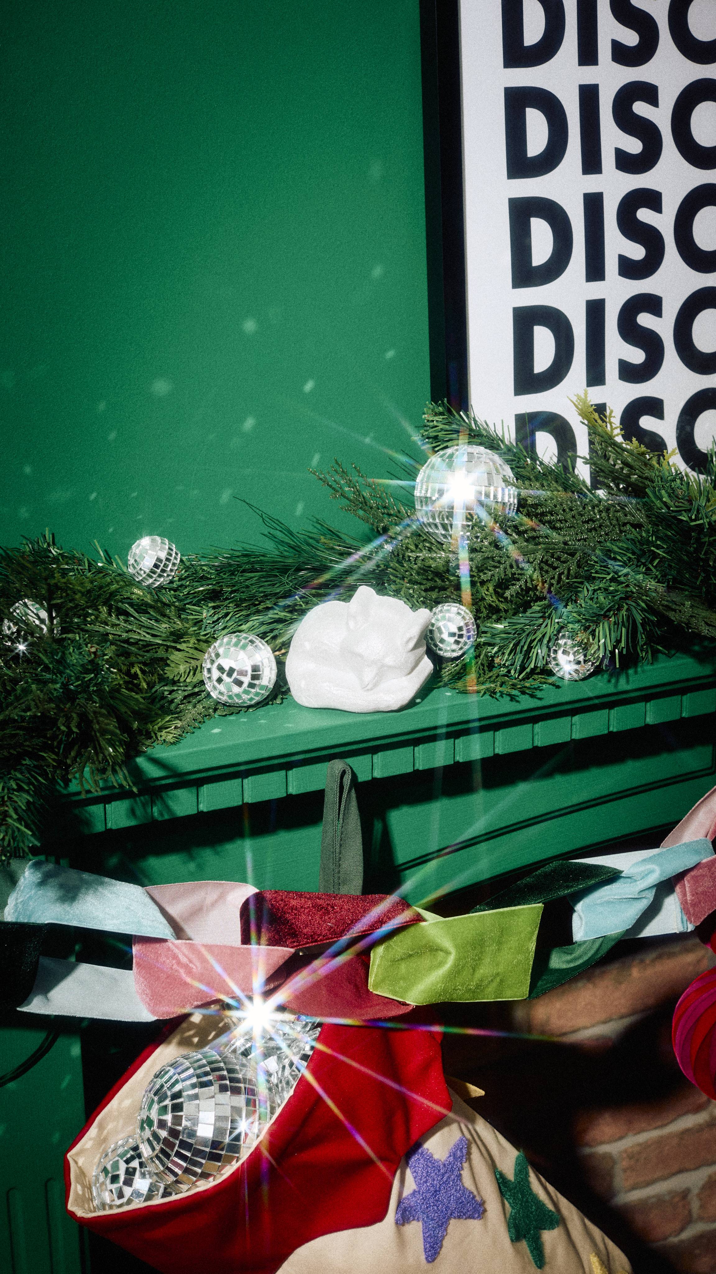 A white, sleeping Arctic fox-shaped bath product rests among small disco ball baubles on a pine garland along a green mantelpiece, above a colourful velvet ribbon garland and a stocking stuffed with mirrored spheres.