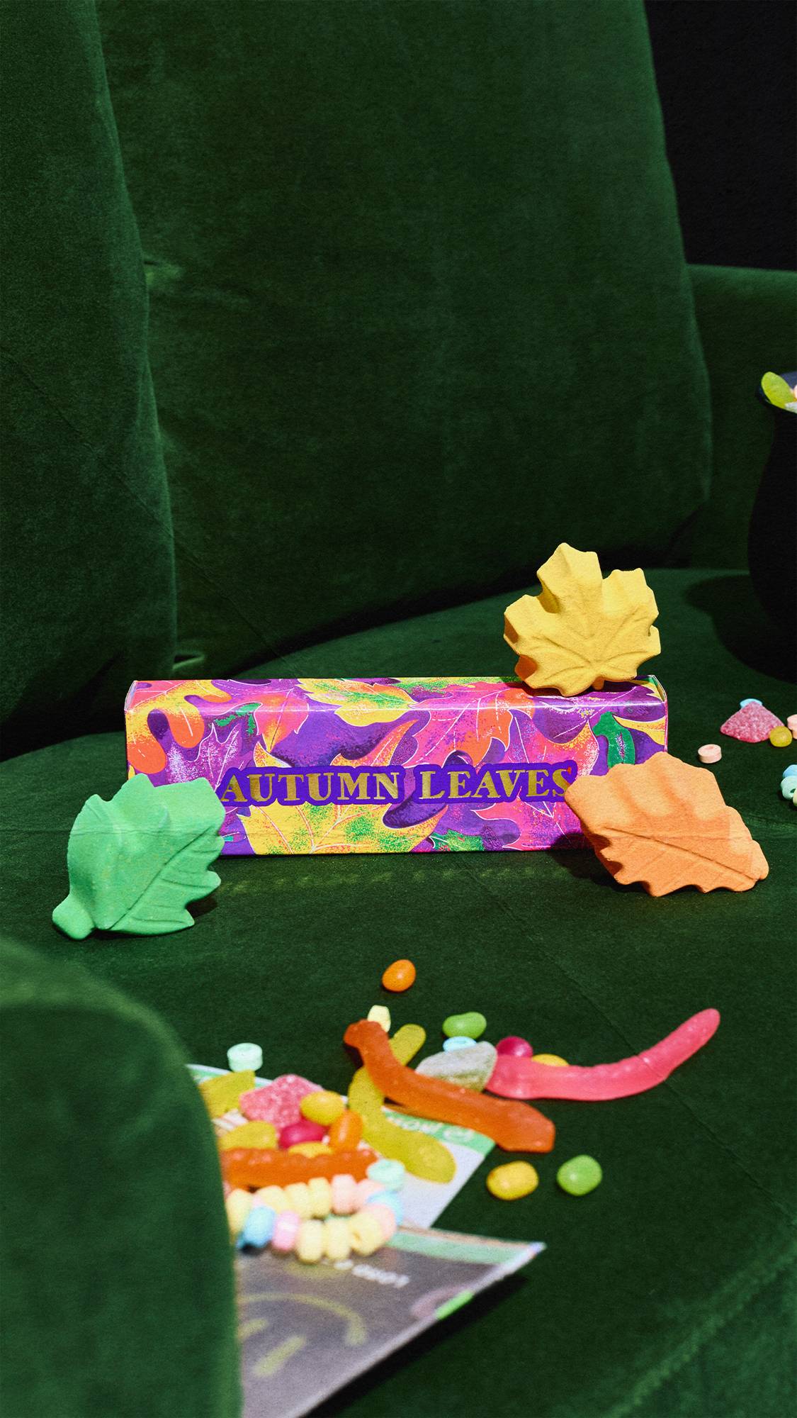 Green, yellow, and orange leaf-shaped bath bombs are displayed around a colourful "Autumn Leaves" box on a plush green velvet armchair, surrounded by scattered sweets like jelly worms and candy necklaces.
