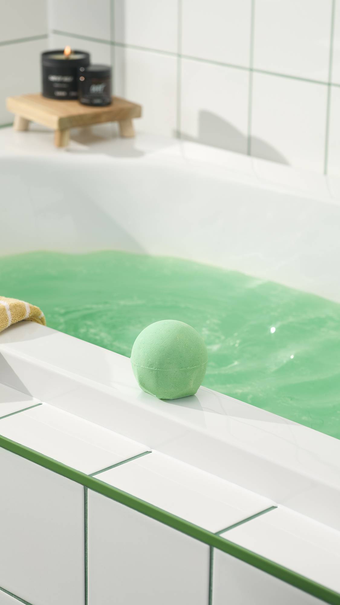 The green Avobath Bath bomb sits on the white edge of a bathtub filled with shimmering green water. In the background, lit candles rest on a small wooden stand against a white tiled wall.