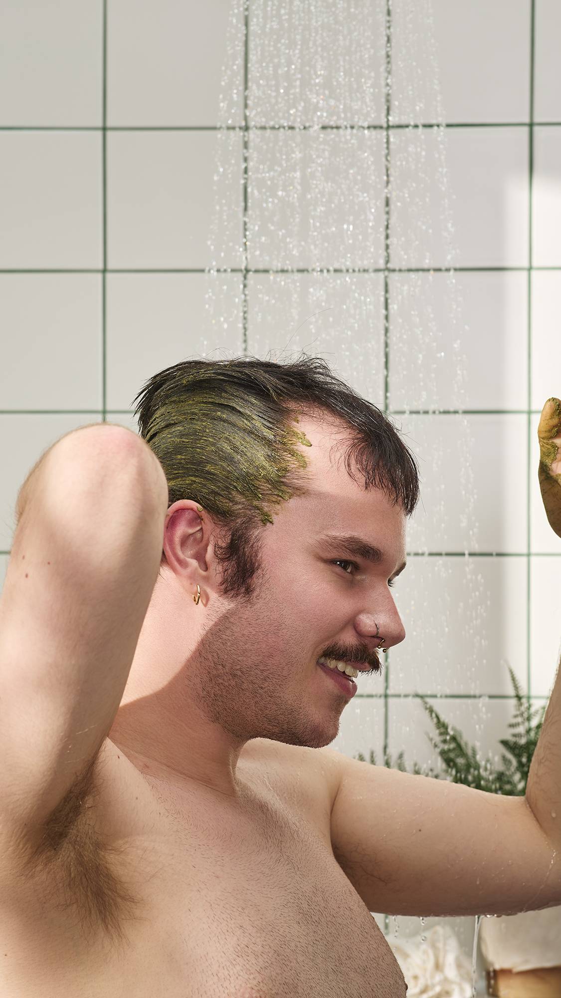 A person with a moustache and short dark hair smiles while applying a thick, olive-green treatment to their hair and scalp in the shower. Water falls in the background against white tiles with green grout, under bright light. 
