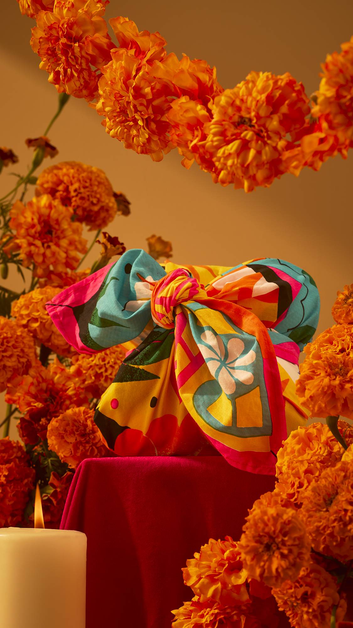 A vibrant, colourful knot-wrapped gift parcel rests on a red pedestal, surrounded by garlands and clusters of bright orange marigolds with a lit candle in warm lighting.