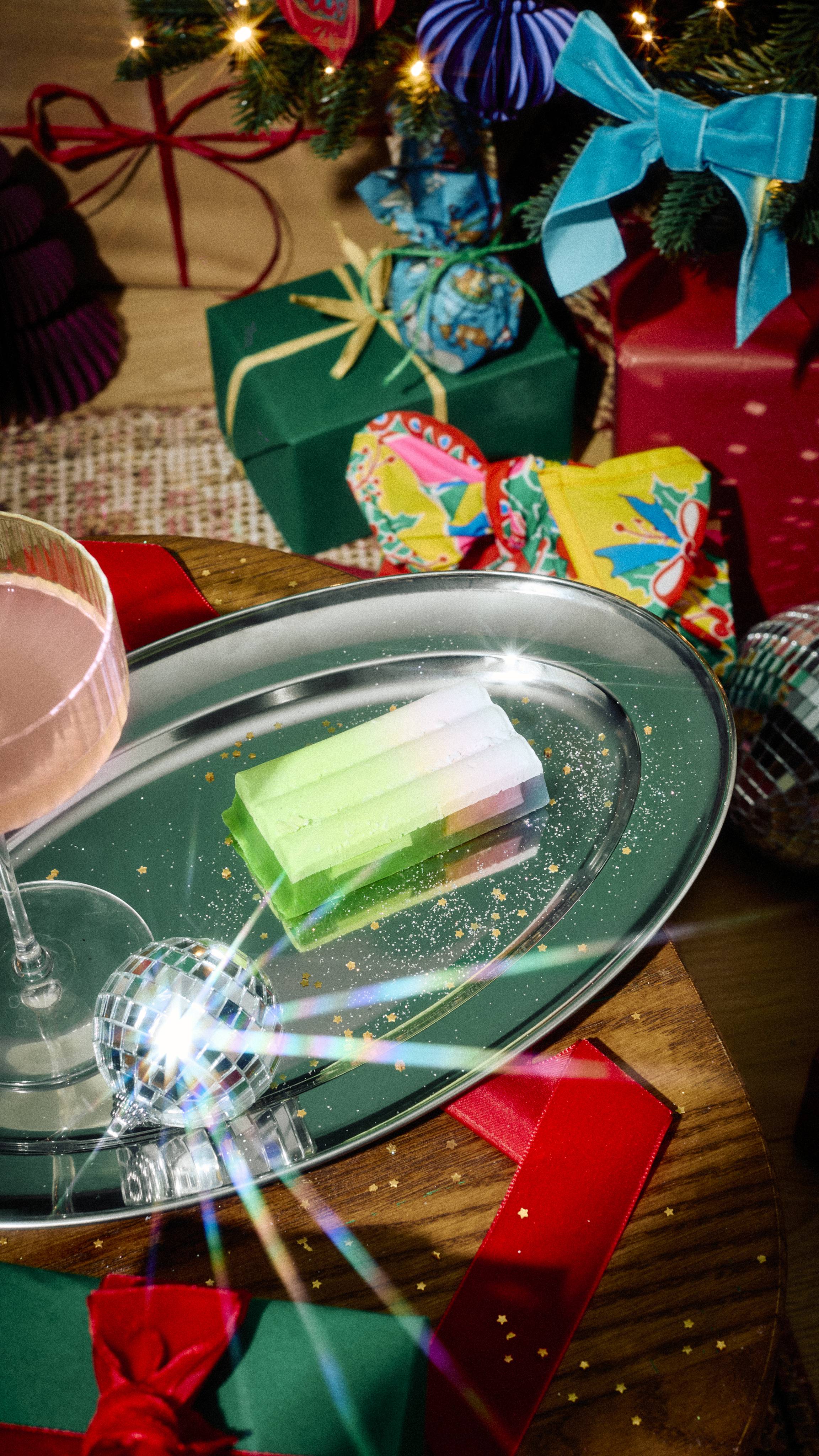 A rectangular green and pastel "Baked Alaska" Lush soap bar rests on an oval silver tray on a wooden table, surrounded by a glass of pink drink and festive wrapped gifts under a decorated Christmas tree.