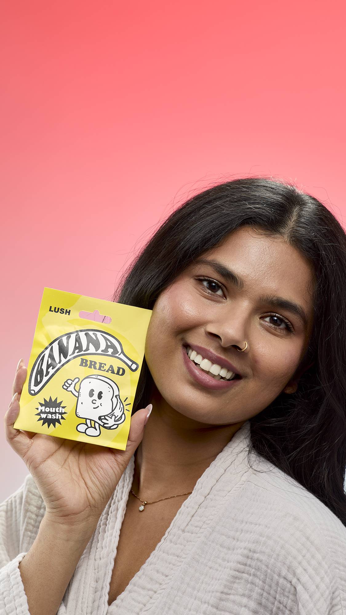 A smiling person with long dark hair holds a bright yellow sachet of Lush Banana Bread mouthwash tabs next to their face. The packet features a cartoon character of a piece of bread holding a banana. 
