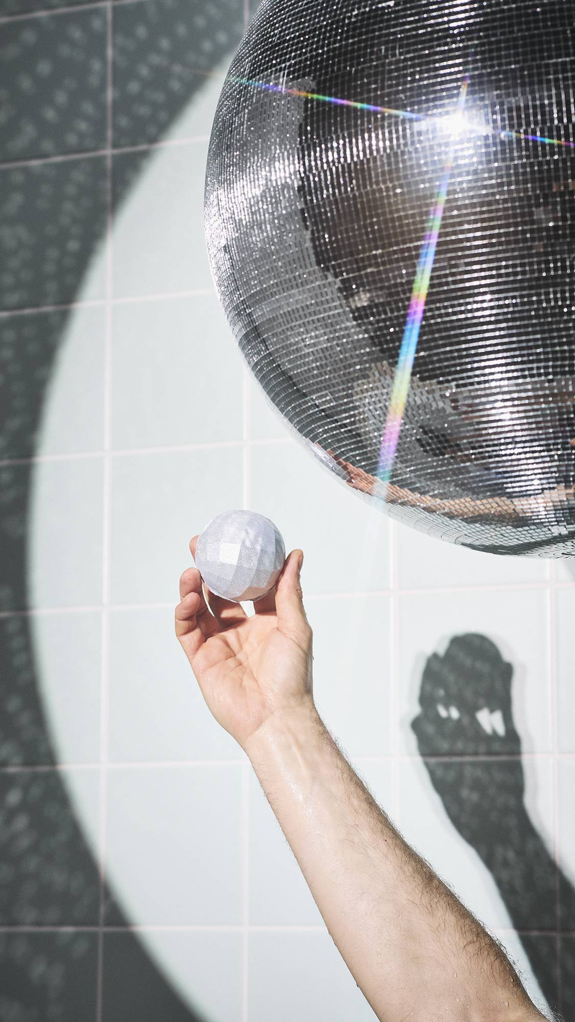 A hand holds a shimmering, "Bathe All The Time" bath bomb with a faceted surface. It is held up towards a large, sparkling disco ball in a bathroom, with a beam of light creating bright reflections and shadows against the pale-tiled wall.