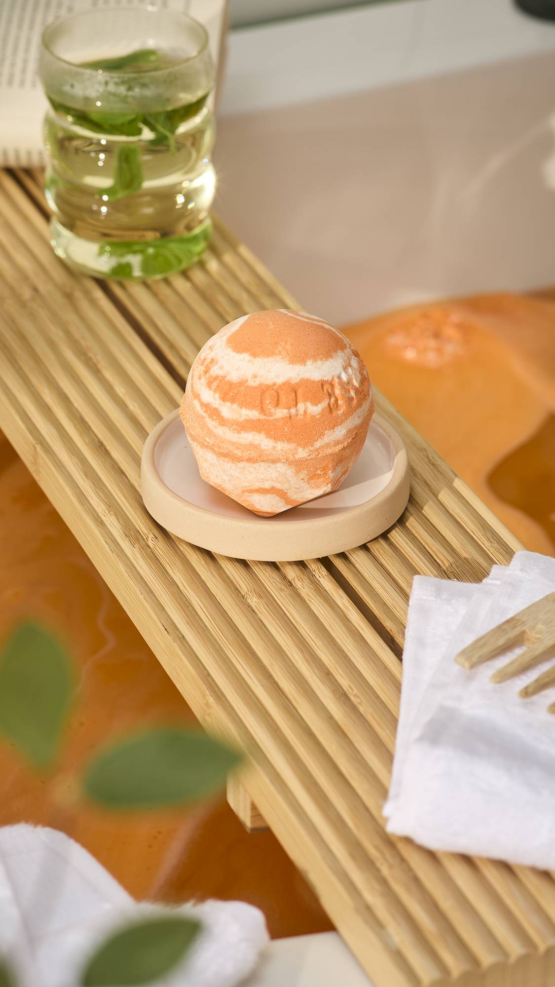 A LUSH "Bather's Digest" bath bomb with orange and white marbled swirls sits on a small dish atop a slatted wooden bath caddy, alongside a glass of iced tea, with warm orange bathwater visible below.