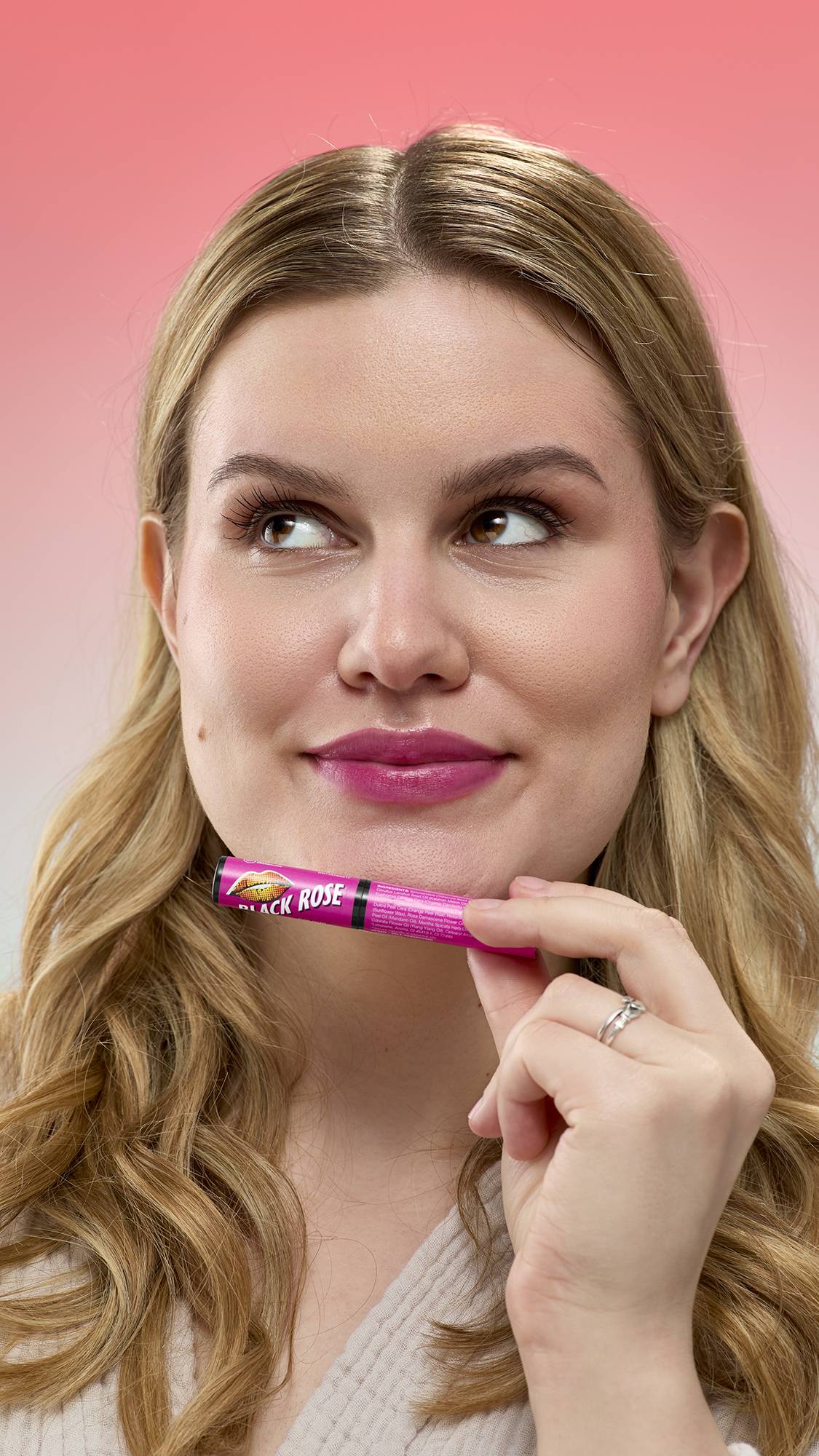 A person with blonde wavy hair looks upward while holding a pink tube of Black Rose Lip Stain under their chin. Their lips are tinted a deep berry color. They are wearing a light robe against a soft pink gradient background.
