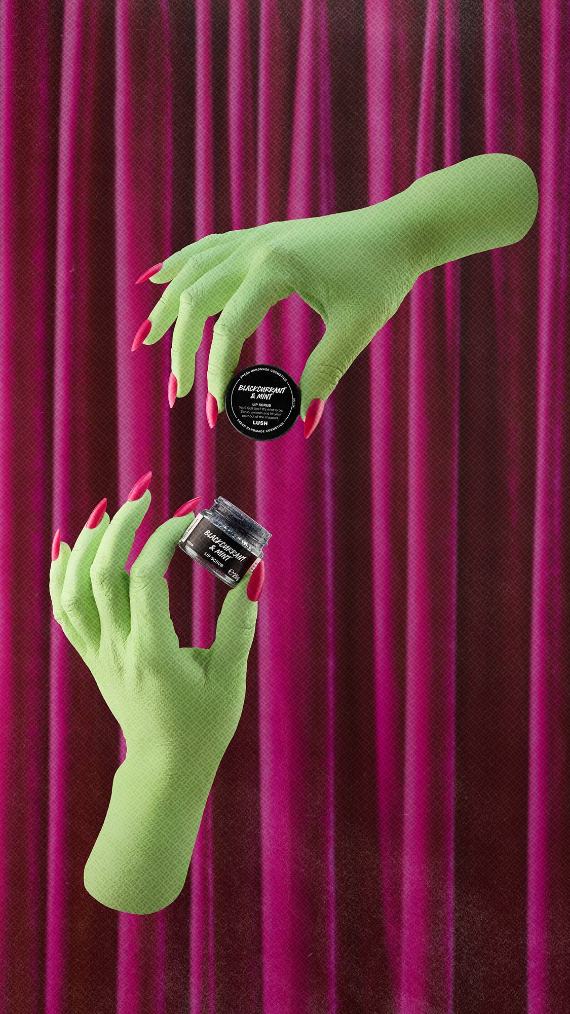 Two pale green witch's hands with long pink nails are positioned against deep pink velvet curtains. One hand holds the black lid and the other holds an open small pot of Lush 'BLACKCURRANT & MINT' lip scrub, filled with a dark textured scrub.