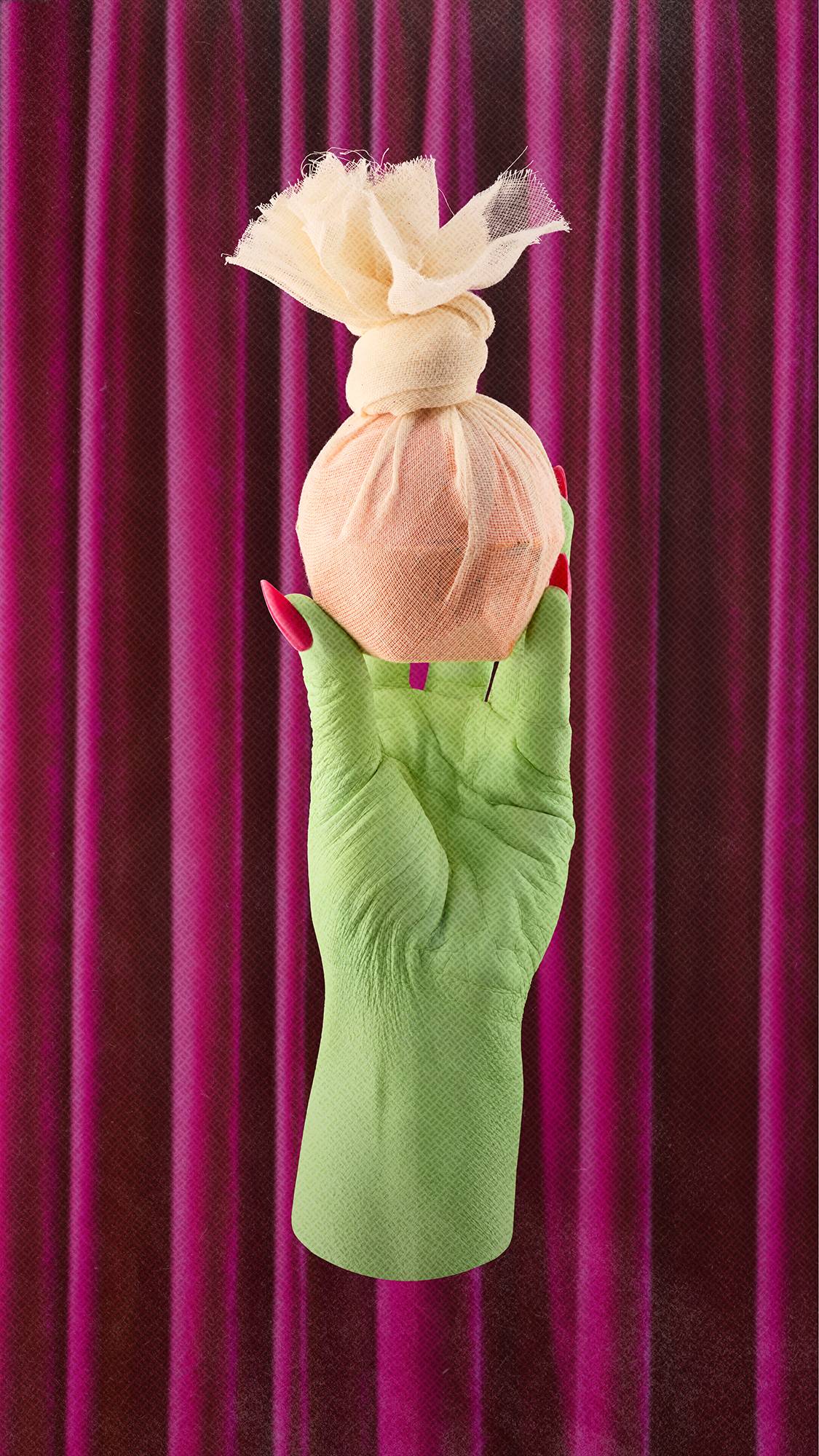 A green hand with long, bright pink nails holds up a round bath bomb wrapped in cream-coloured muslin cloth, tied at the top. The hand reaches upward against a backdrop of rich, pleated magenta velvet curtains.