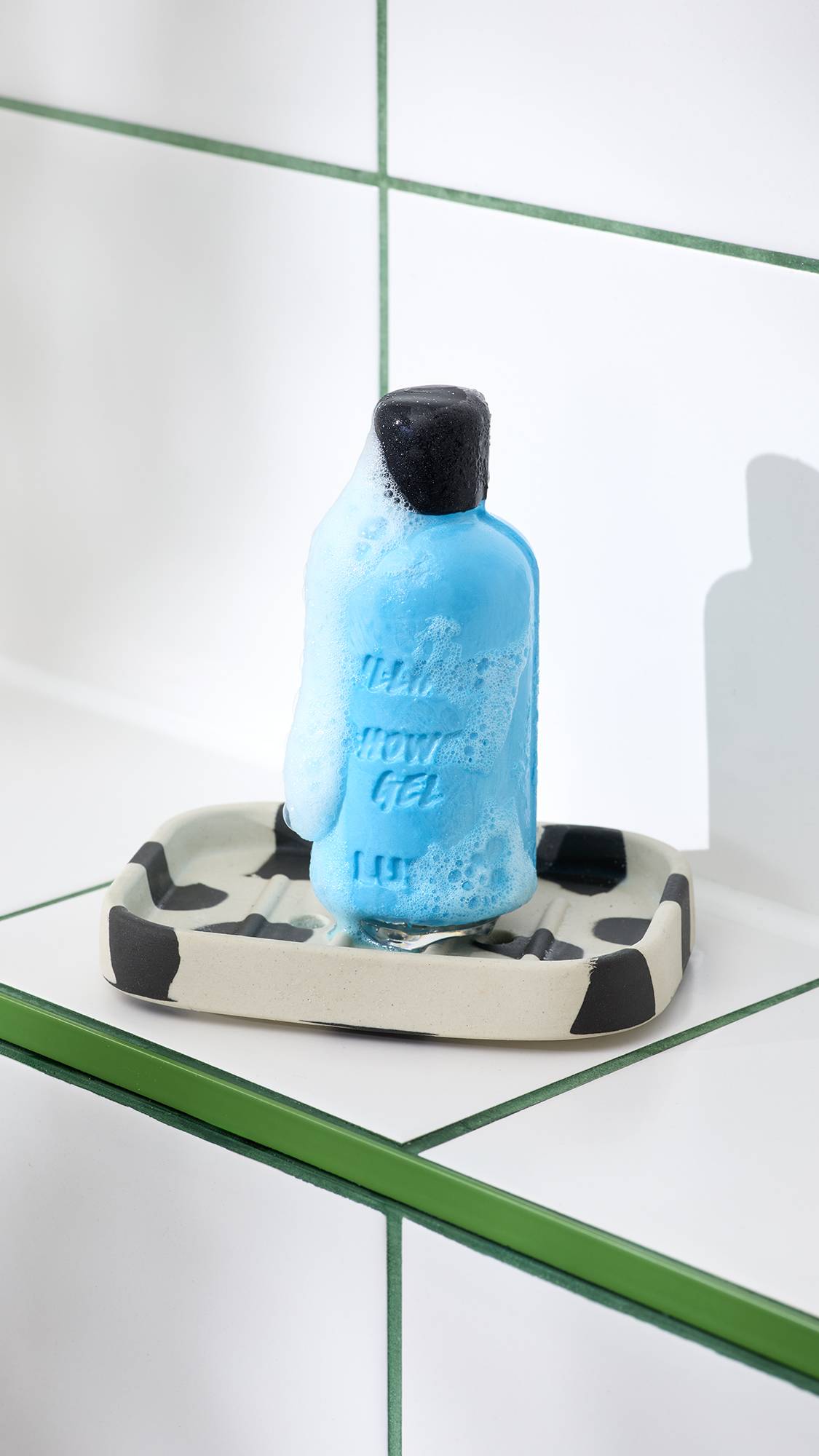 A bright blue solid shower gel bar shaped like a bottle rests on a black and cream spotted soap dish, partially covered in frothy foam. It sits against a background of white tiles with green grouting.