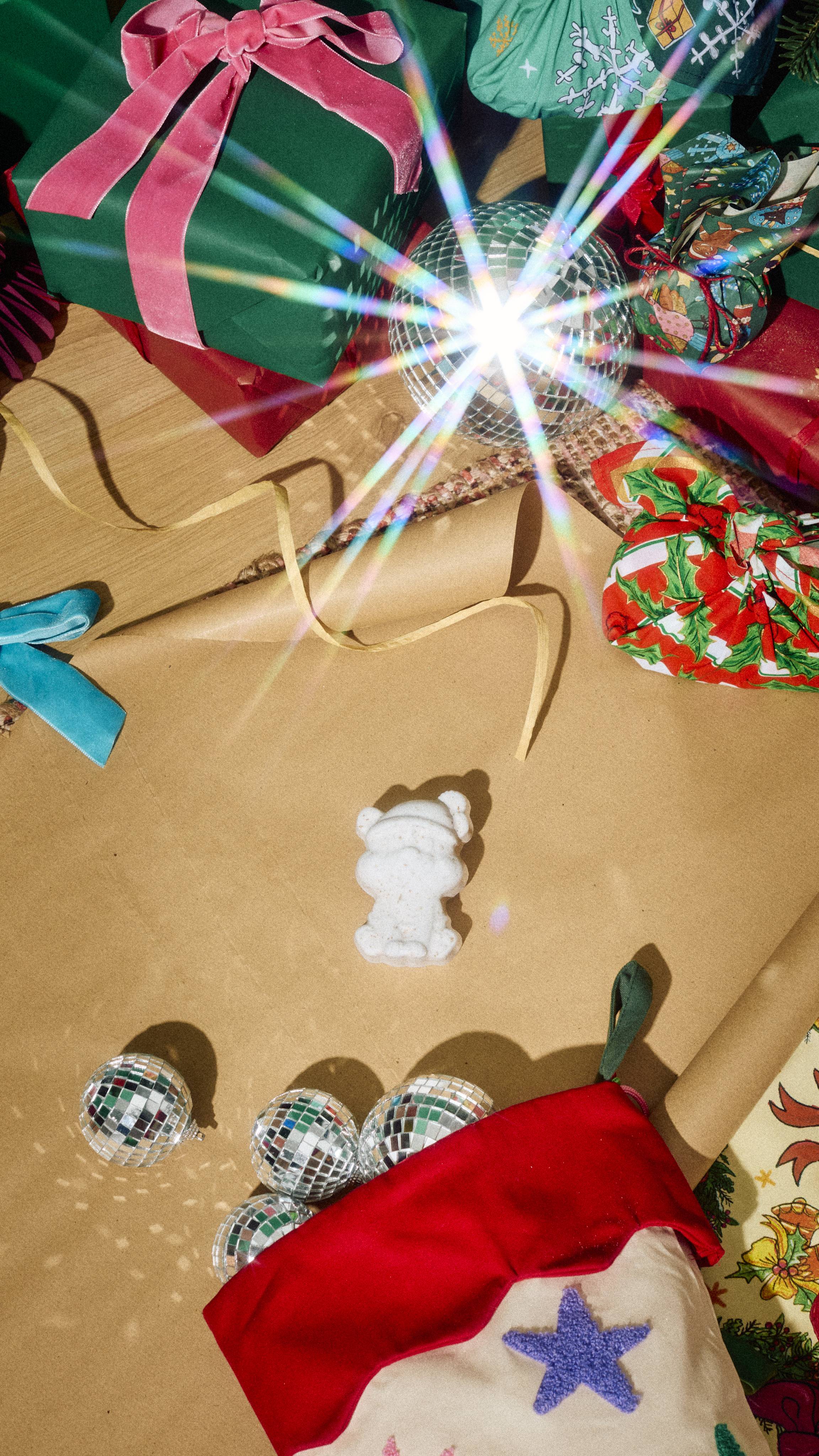 A white Lush "Butterbear" bath bomb sits on brown wrapping paper alongside a stocking filled with miniature disco balls, surrounded by green wrapped gifts tied with pink ribbon and sparkling light reflections.