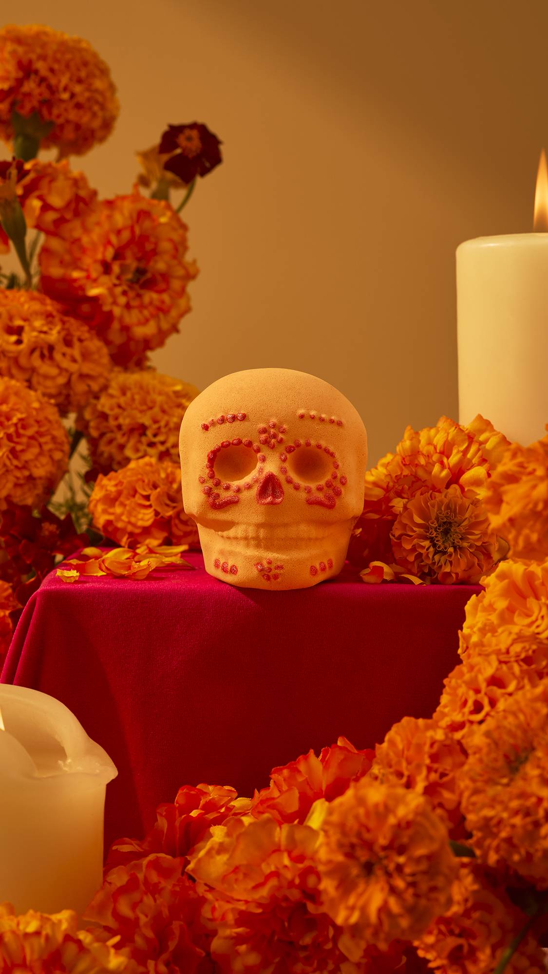 An orange sugar skull-shaped bath bomb sits on a red cloth-covered table surrounded by vibrant orange marigold flowers and lit candles in a warm, festive display.
