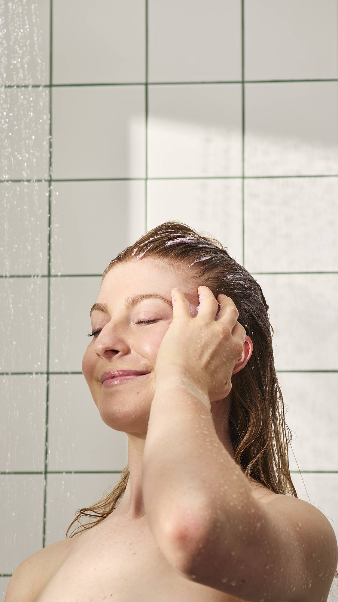 A model with long, reddish-brown hair smiles with closed eyes while massaging a lilac-coloured product into their scalp in the shower. Water falls in the background against white tiles with green grout, illuminated by soft sunlight.