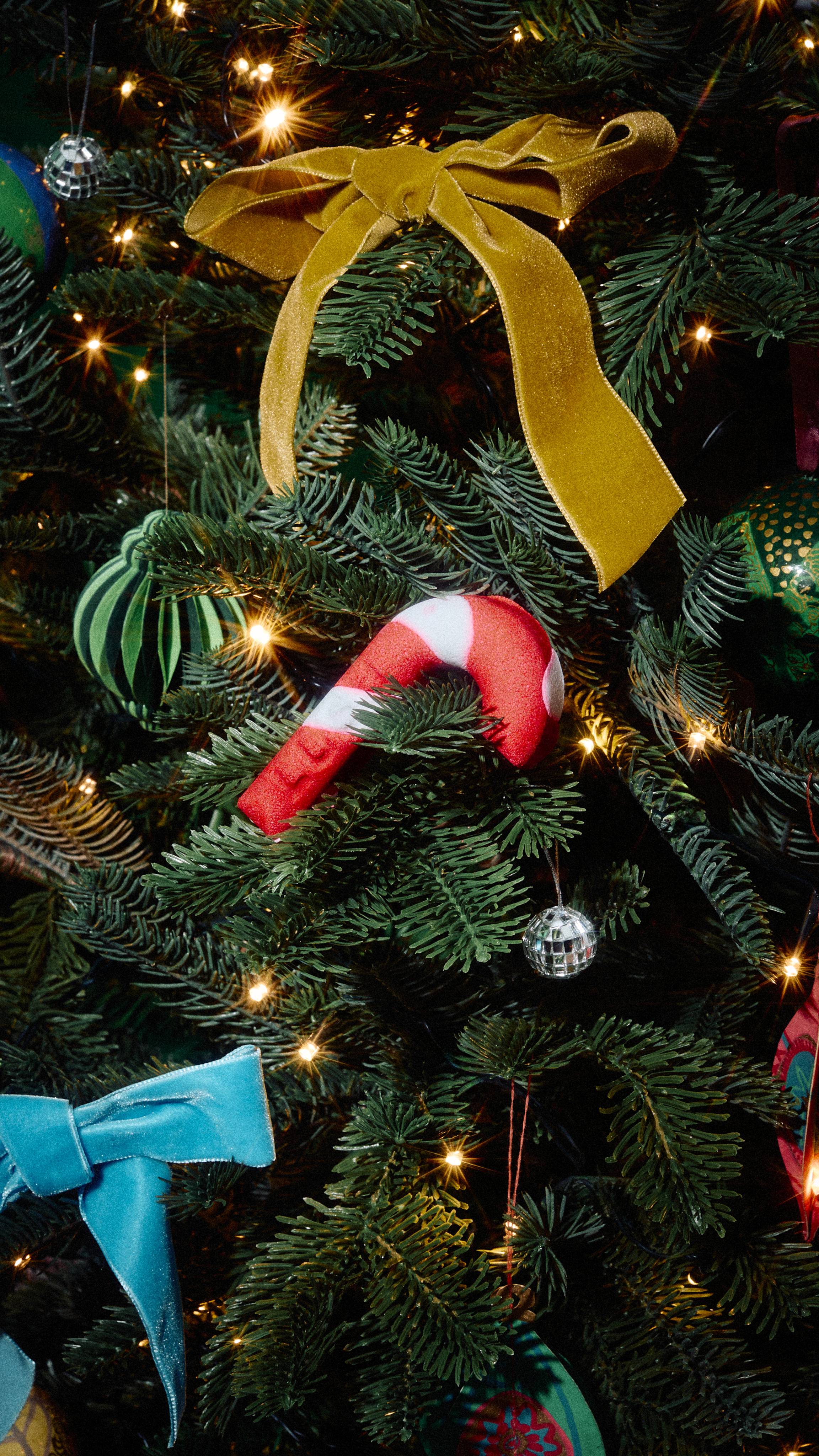 A red and white striped candy cane bubble bar nestles on a branch of a decorated Christmas tree, amidst warm fairy lights, a mustard yellow velvet bow, blue ribbons, and tiny disco ball baubles.