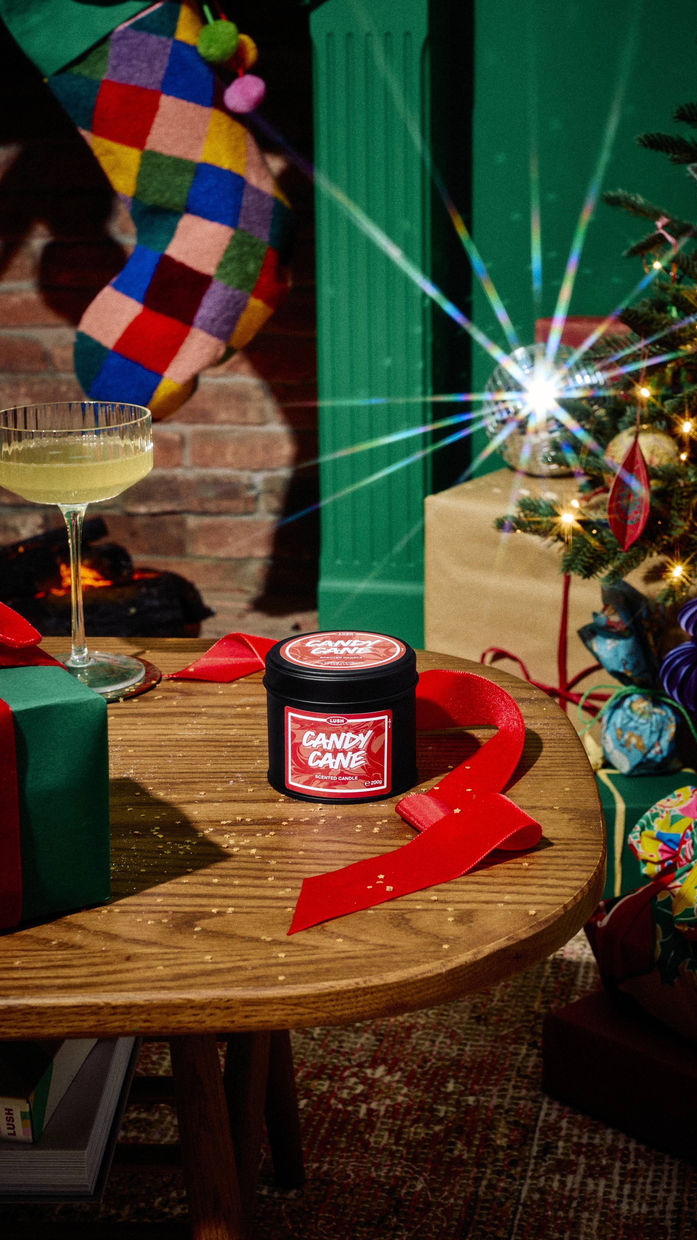 A black tin of Lush Candy Cane scented candle sits on a wooden table beside curling red ribbon, a cocktail glass, and a wrapped gift, set in a festive room with a Christmas tree and colourful stocking.