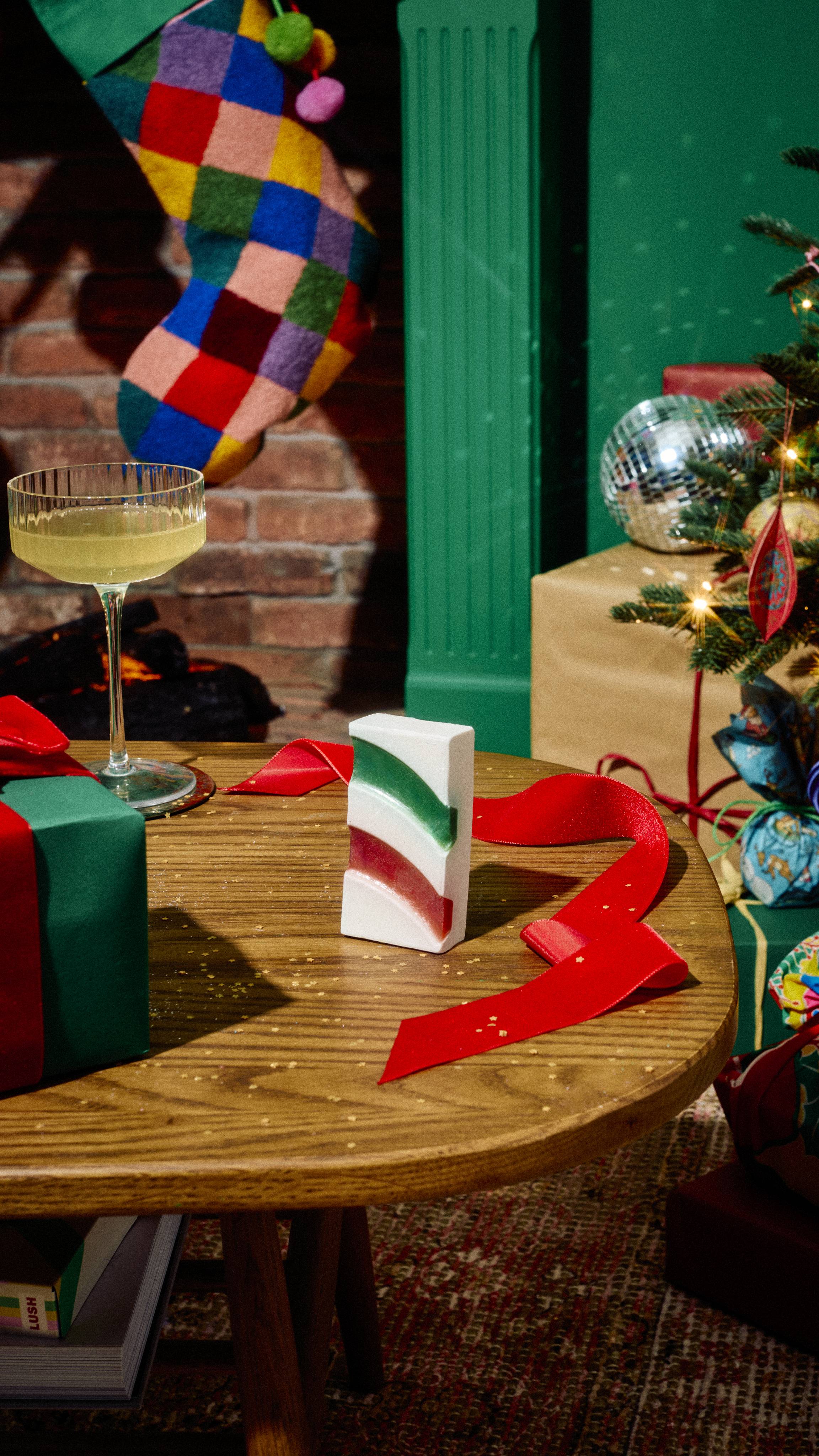 A rectangular white "Candy Cane" soap with green and red wavy stripes stands upright on a wooden table alongside a cocktail glass, red ribbon, and wrapped presents near a colourful Christmas stocking.