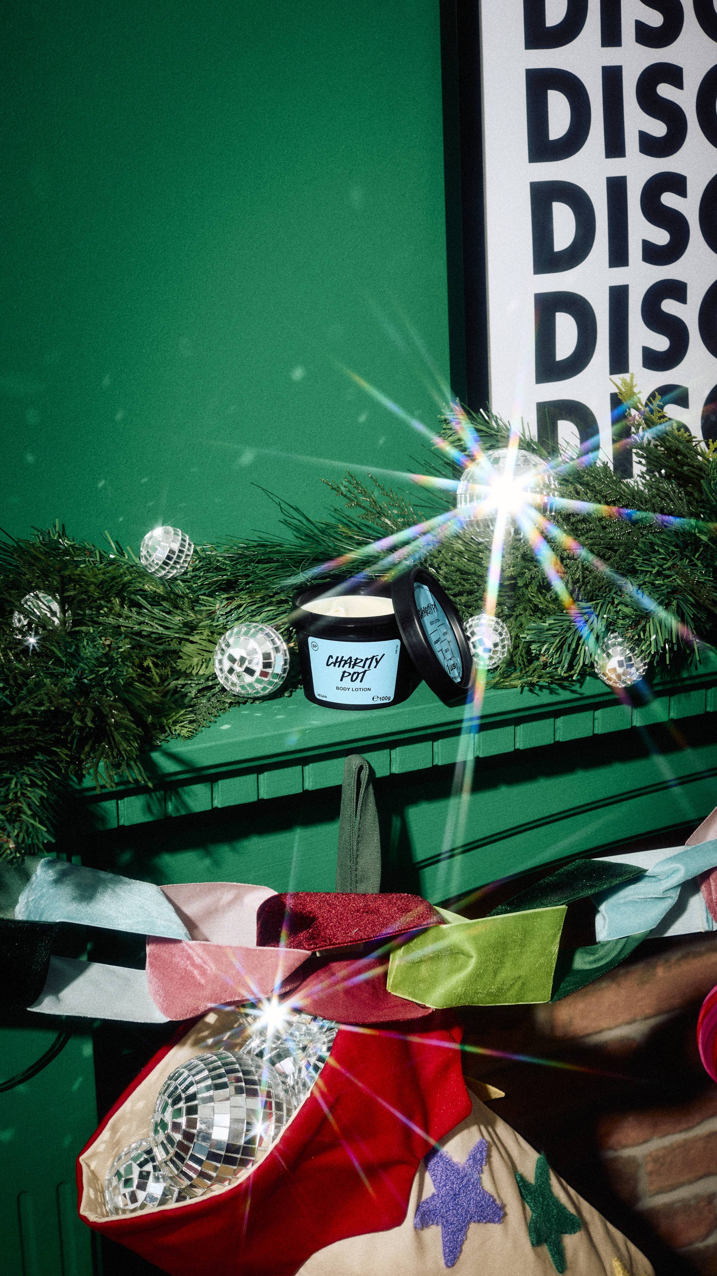 An open black tub of Lush Charity Pot body lotion rests on a green mantelpiece among pine garland and mini disco balls, above a colourful stocking filled with mirrored baubles.