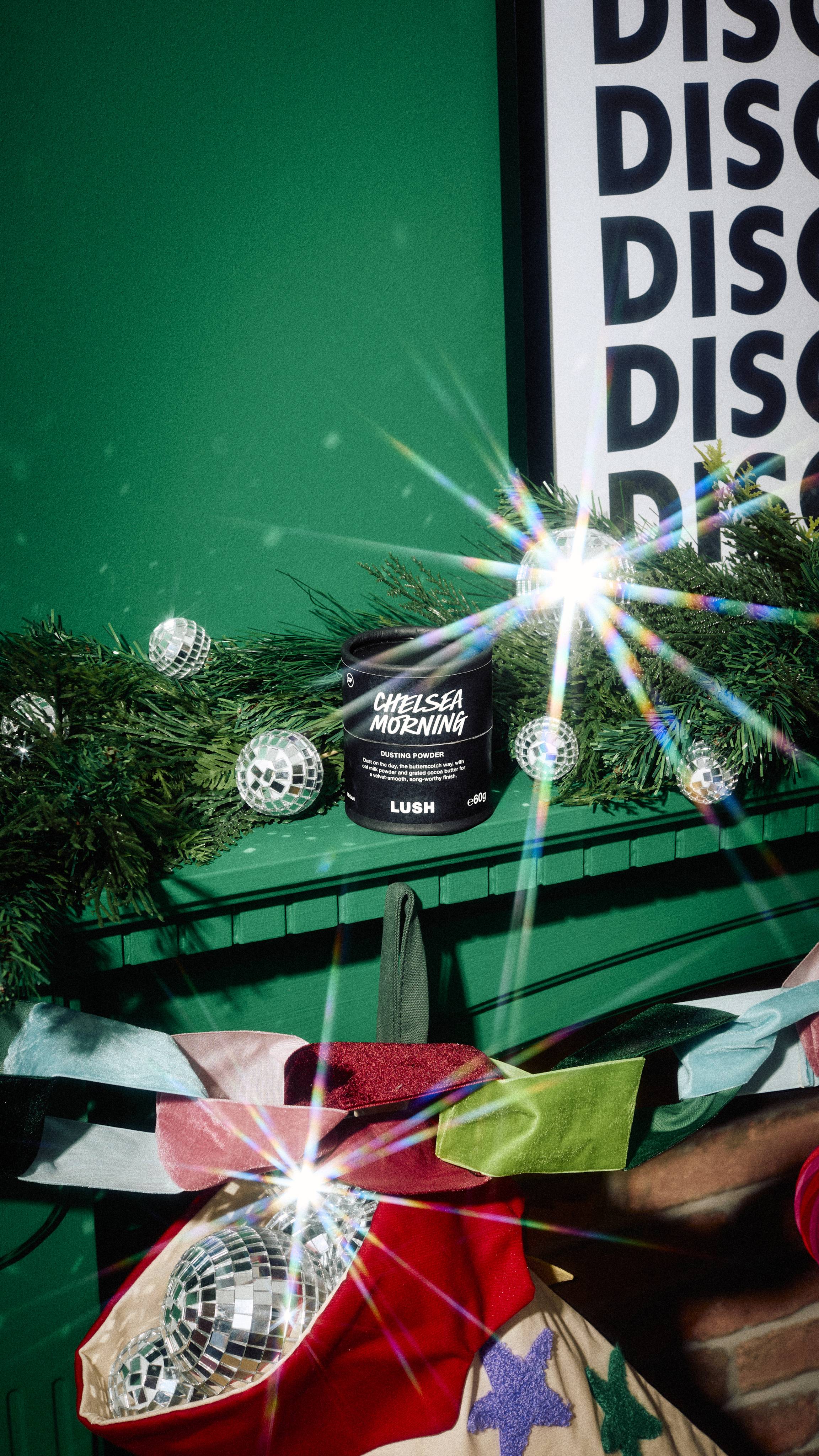 A black card tub of Lush Chelsea Morning dusting powder rests on a green mantelpiece nestled among pine garland and mini disco balls, set below a framed print and above a colourful Christmas stocking.