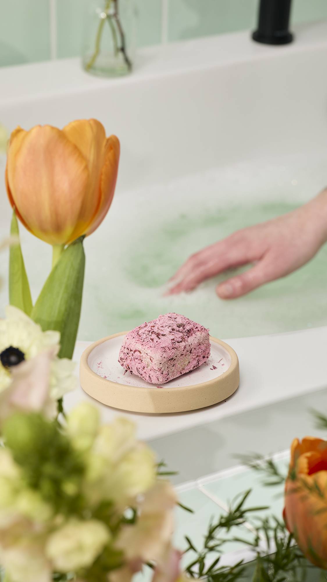 A pink, textured exfoliating bar sits on a small beige dish on a white bath ledge. Blurred orange tulips frame the foreground, while a hand reaches into pale green foamy bathwater in the background.