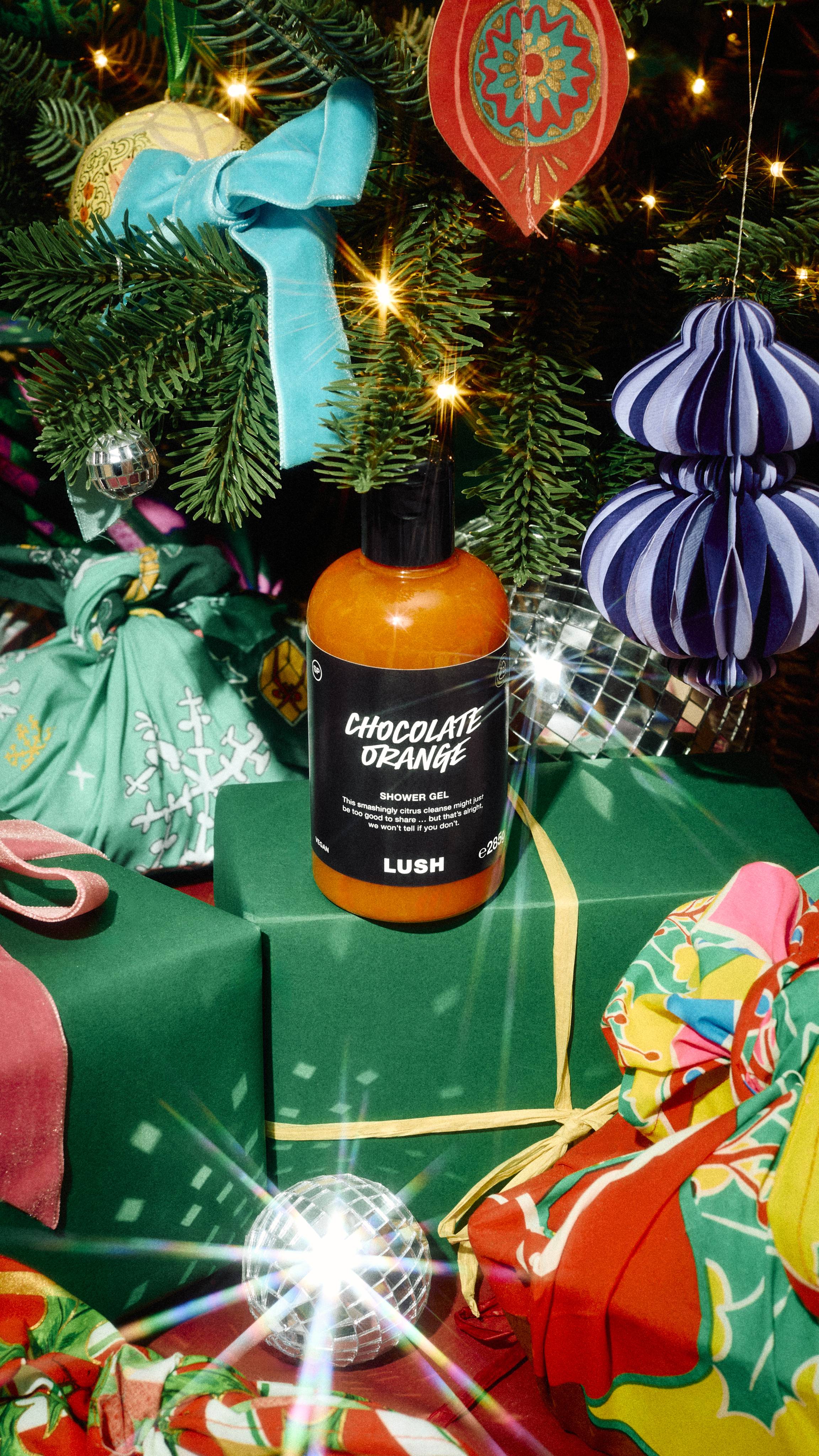 A bottle of Lush "Chocolate Orange" shower gel sits on a green gift box tied with yellow ribbon beneath a decorated Christmas tree, surrounded by colourful wrapped presents, disco balls, and festive baubles.