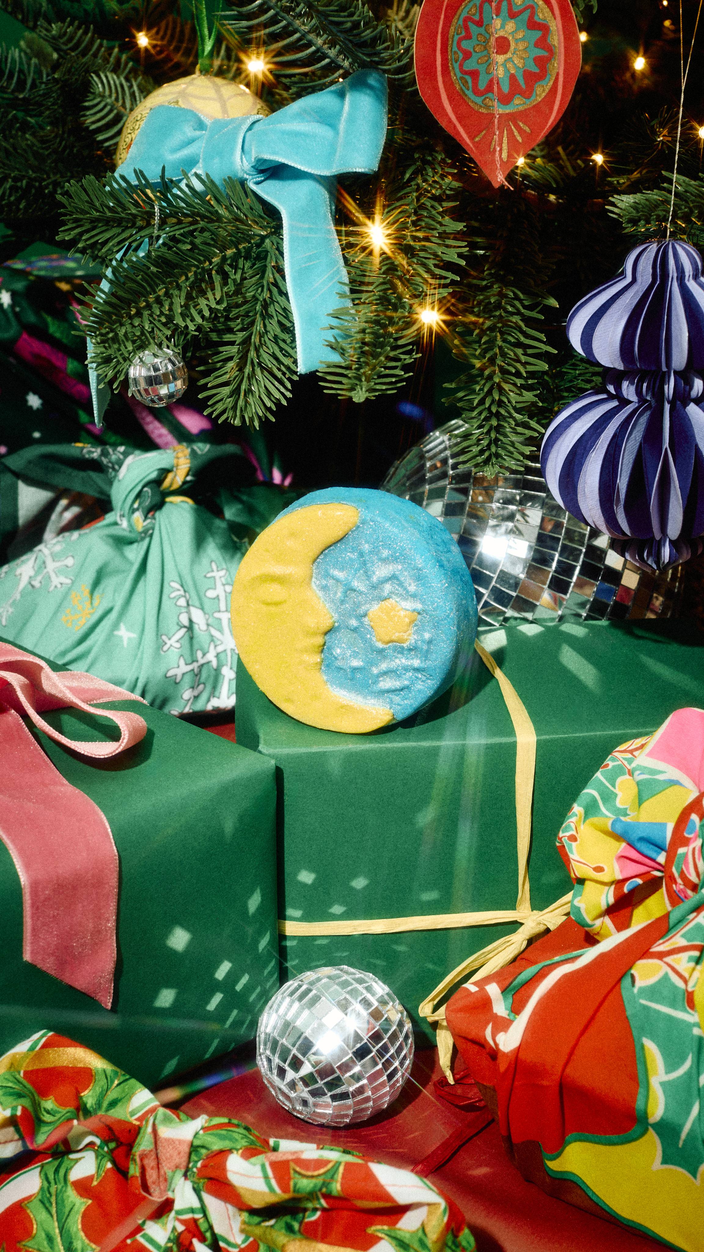 A round yellow and blue crescent moon bubble bar rests on a green wrapped present beneath a decorated Christmas tree, surrounded by disco balls, colourful bows, and patterned gift wrap.