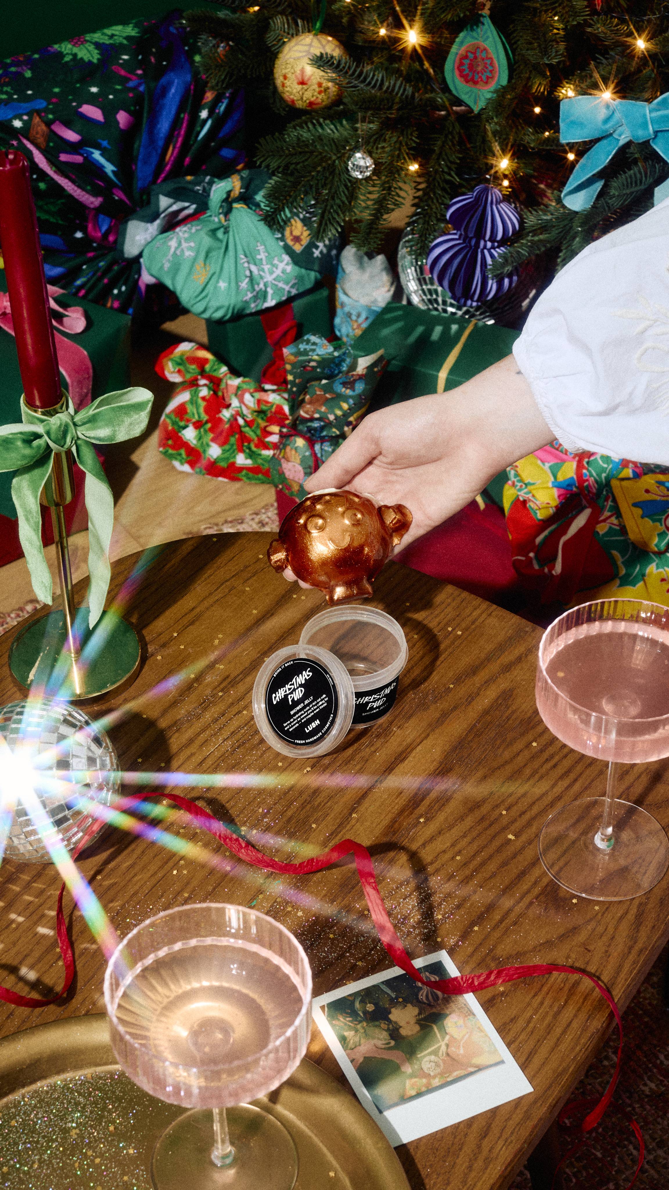 A hand holds a bronze Christmas-pudding-shaped shower jelly over its clear tub labelled "Christmas Pud" on a wooden table, surrounded by pink drinks, red ribbon, and festive gifts beneath a decorated tree.