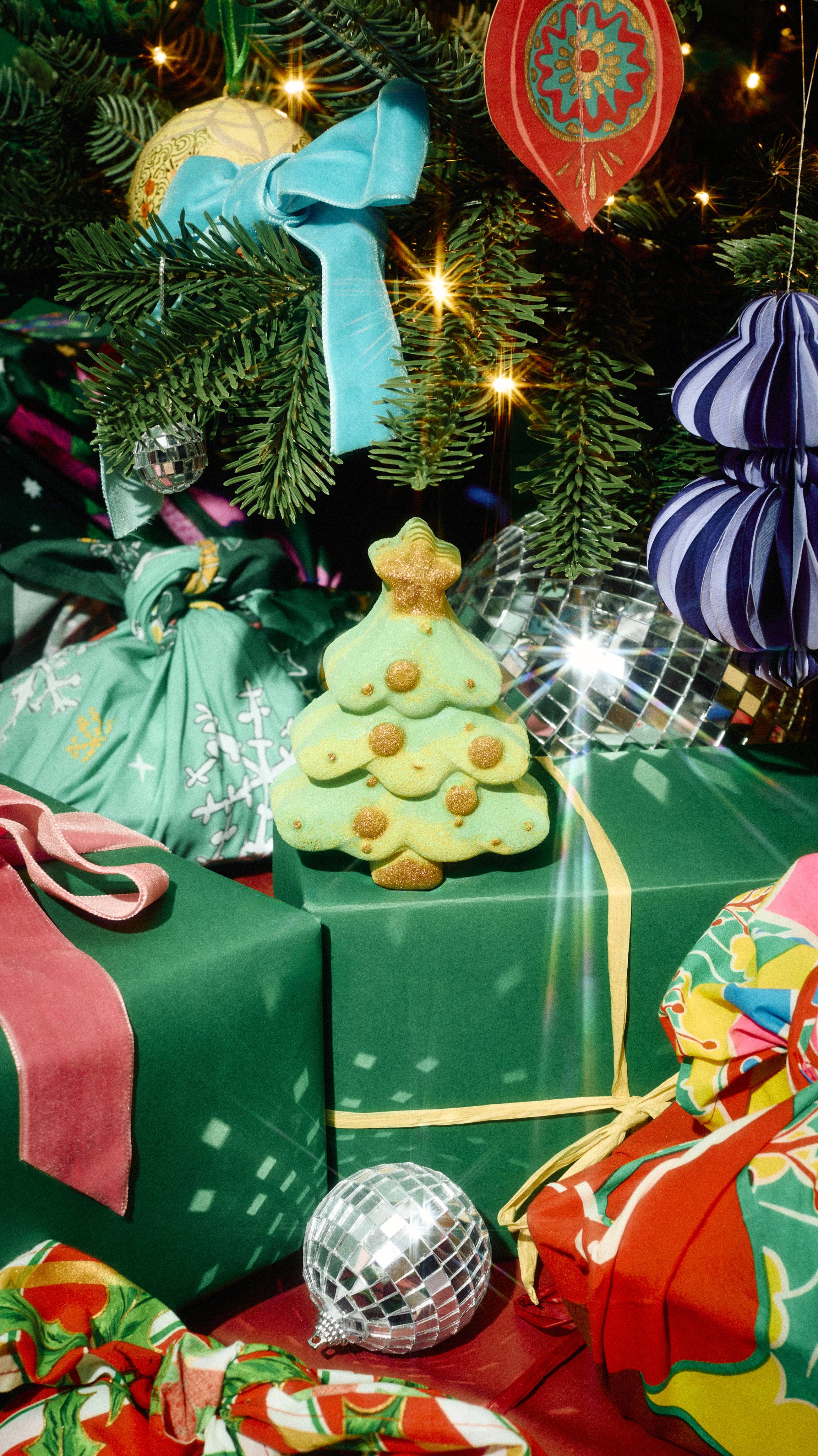 A green Christmas tree bath bomb with golden details sits on wrapped presents under a decorated Christmas tree with blue bows and disco ball baubles.