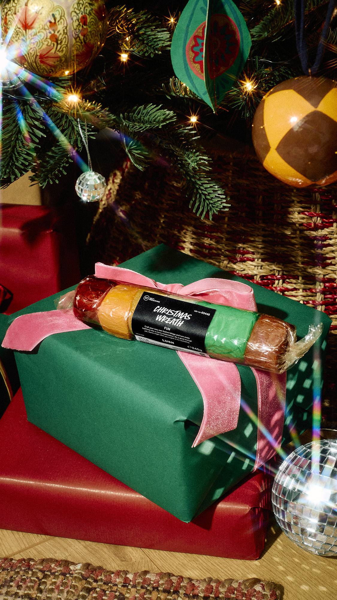 A wrapped Lush "Christmas Wreath" FUN multi-coloured mouldable bath soap bar sits on a green gift box tied with a pink velvet bow beneath a twinkling Christmas tree surrounded by festive ornaments.