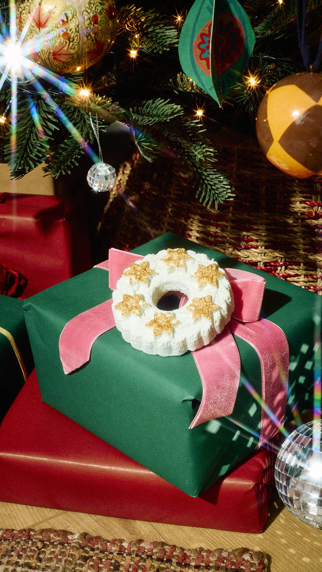 A white wreath-shaped bubble bar topped with gold stars rests on a green wrapped present tied with a pink velvet bow beneath a twinkling Christmas tree surrounded by festive ornaments and gift boxes.