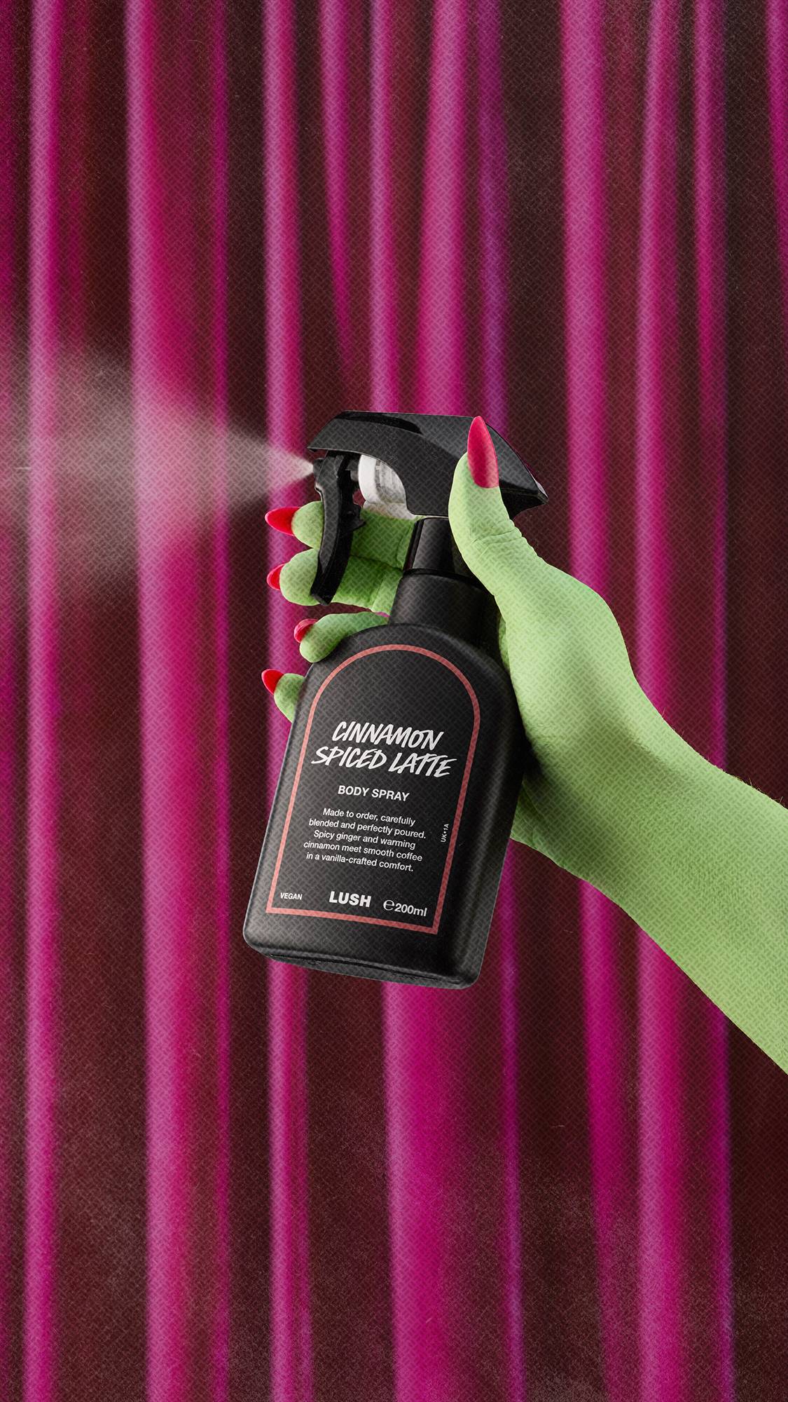 A green-skinned hand with bright pink nails sprays Lush Cinnamon Spiced Latte body spray, releasing a fine mist against a backdrop of rich, draped pink curtains.