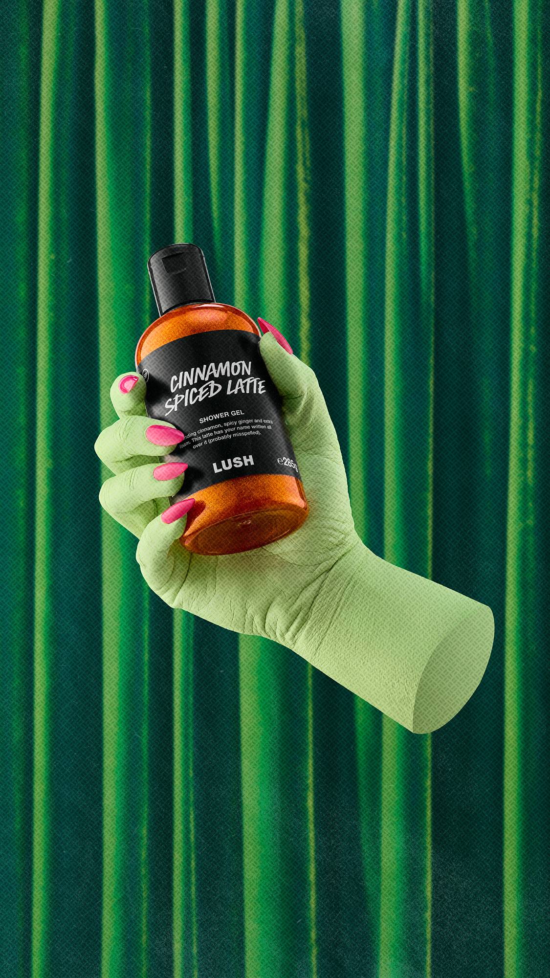 A green hand with bright pink nails holds a bottle of shimmering orange Cinnamon Spiced Latte shower gel against a backdrop of draped green velvet curtains.