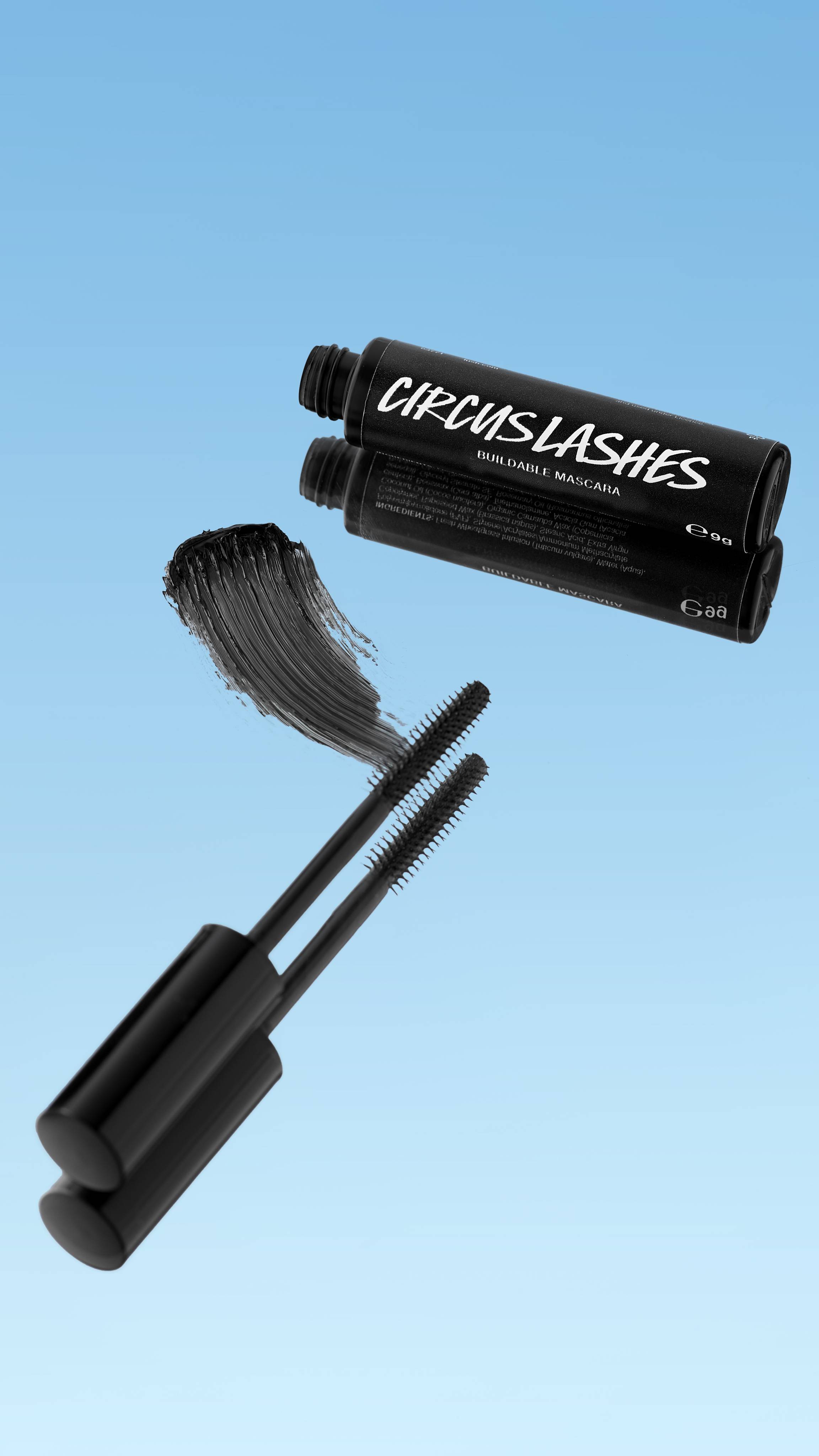 The black tube of Circus Lashes ~ Mascara is shown lying down with its reflection visible. Two black mascara wands are extended below, next to a thick smear of black mascara. The products float against a bright blue background.