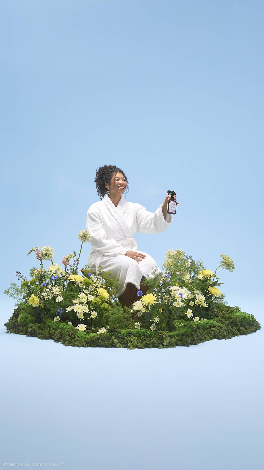 A person with curly hair wearing a white dressing gown sits on a patch of green moss and vibrant wildflowers. They are holding up a black spray bottle of "Cloudberry" body spray, all set against a solid light blue background.