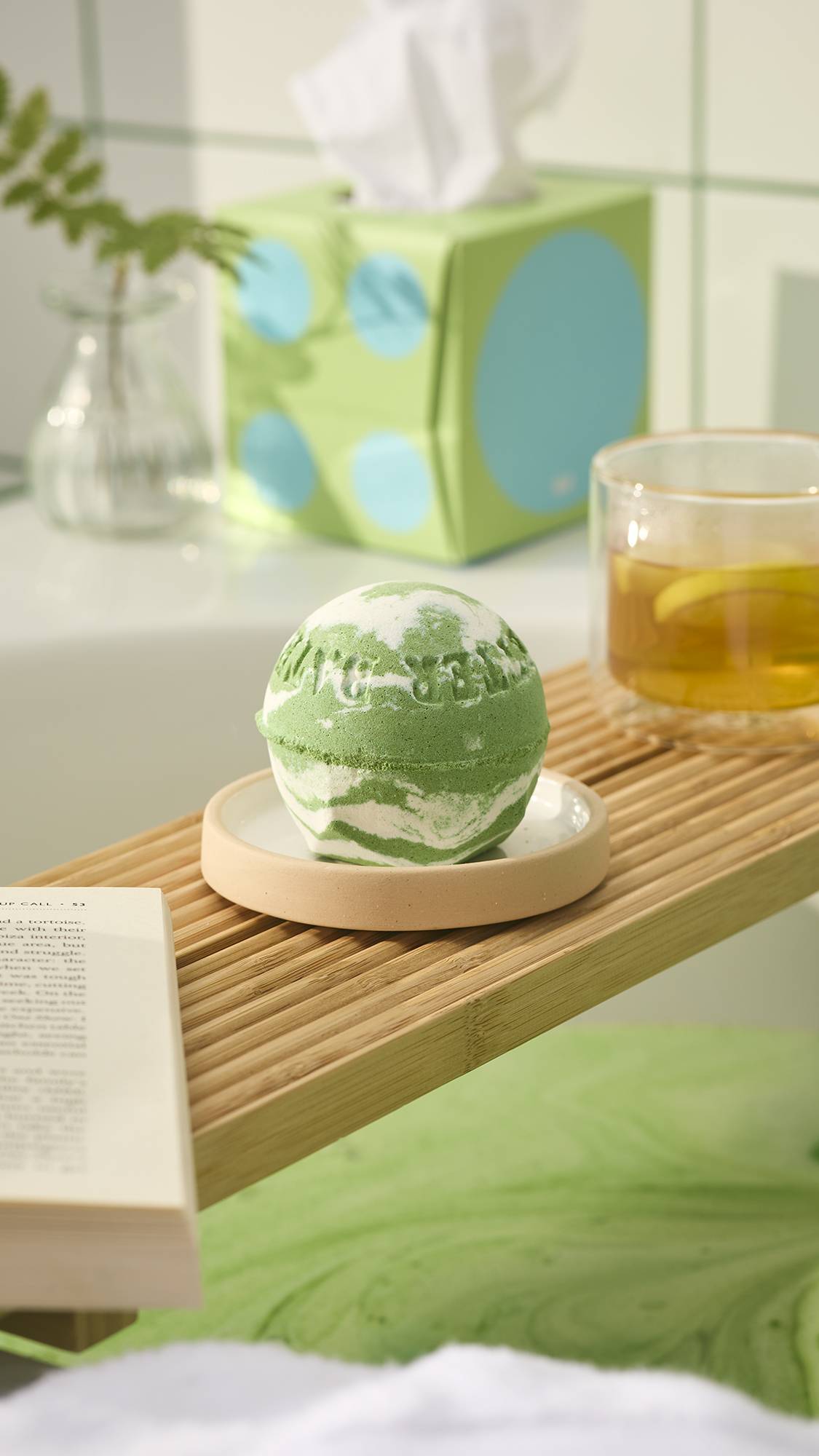 The green and white marbled "Cold Bath Bomb" rests on a wooden bath tray alongside a cup of tea. Green bath water is visible below, framed by mint green tiles and a white towel in the foreground.