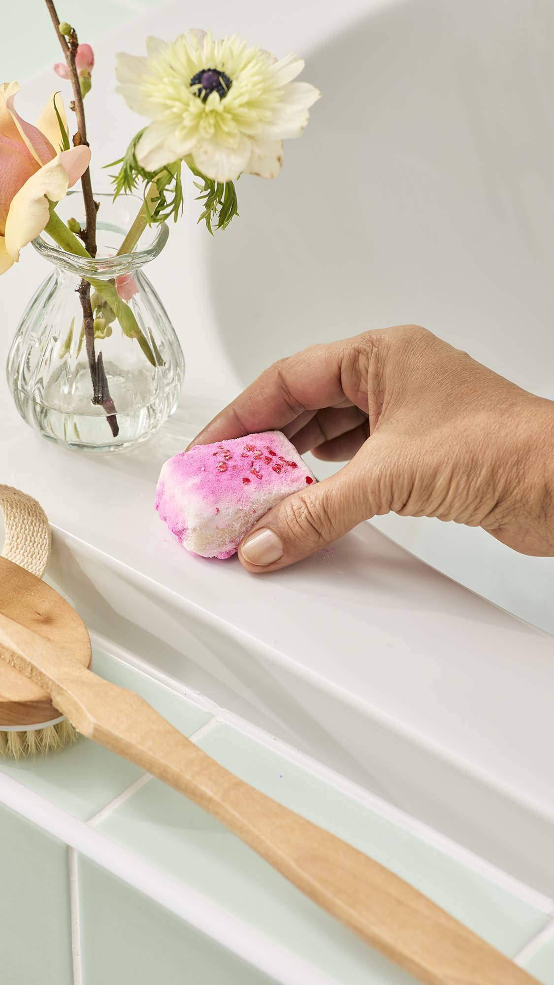 A hand places a small, square "Creamy Candy" bath melt onto a white bath ledge. The melt features pink and white layers topped with small red sprinkles, and sits next to a glass vase of flowers and a wooden bath brush against a mint green tiled wall.