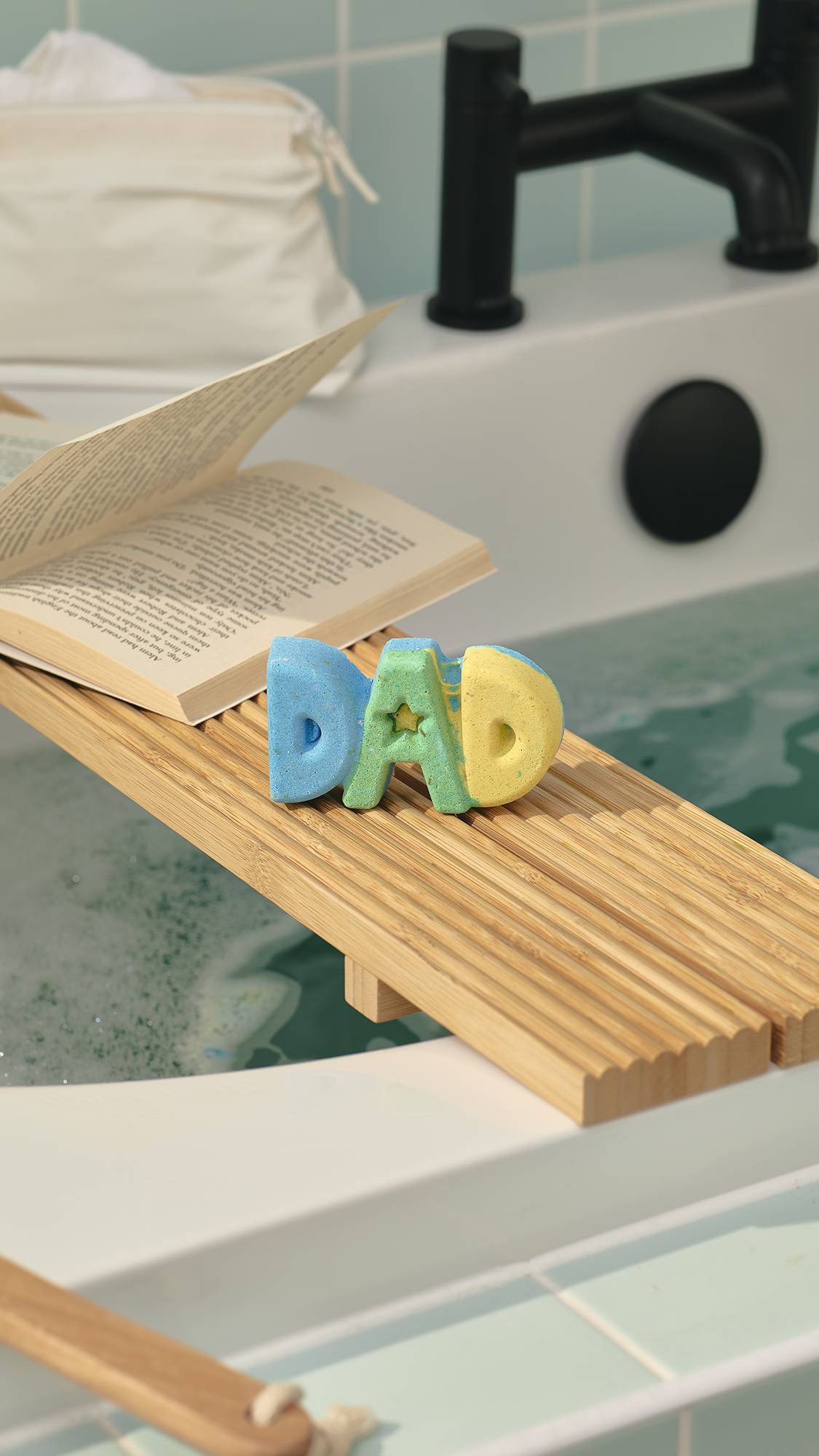 A "DAD" bath bomb in blue, green, and yellow sits on a wooden bath tray over a tub of bubbly bath water. An open book rests nearby. In the background, a white bath with a black tap is set against pale green wall tiles.