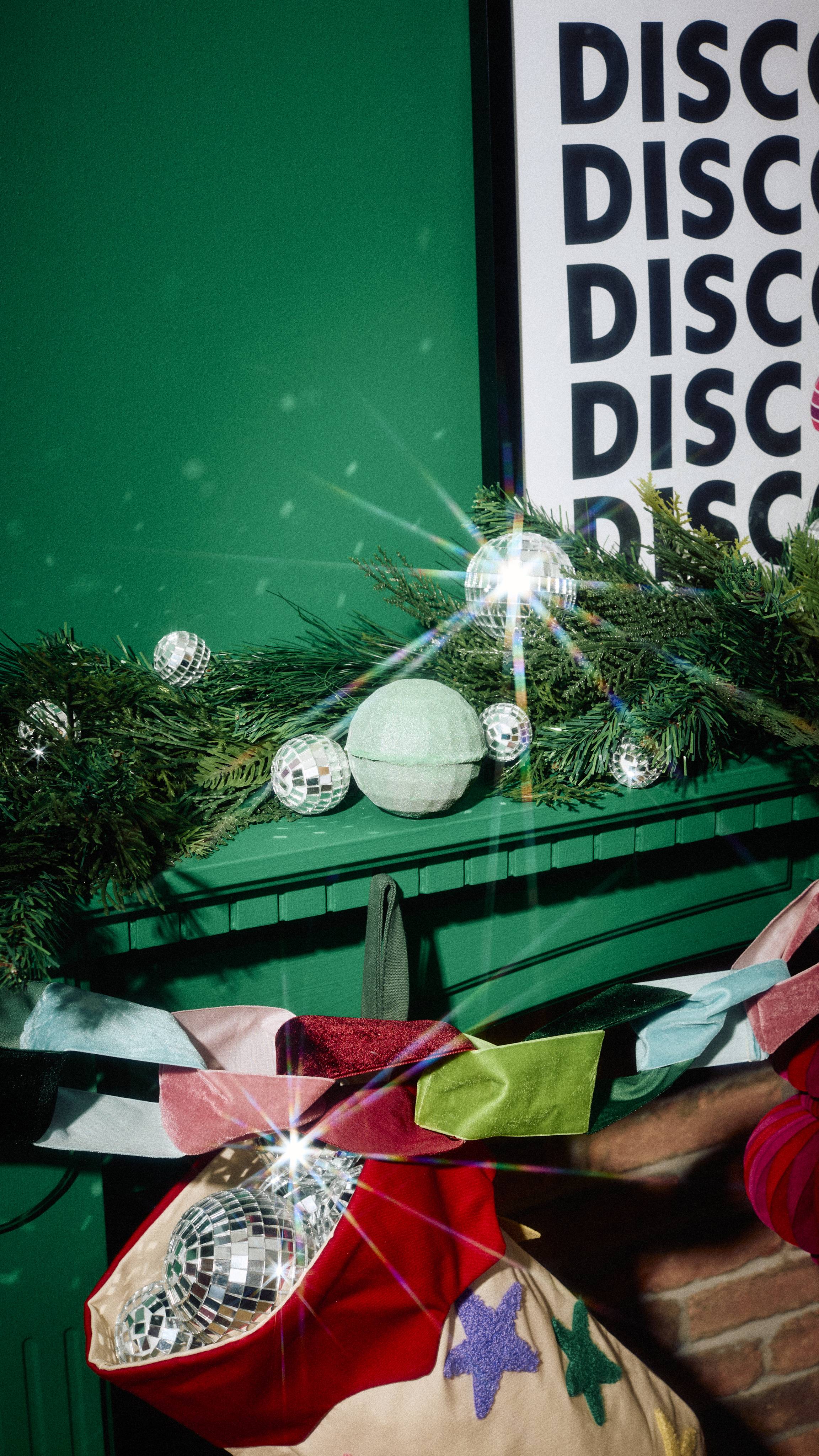 A pale mint-green disco ball-shaped foot bomb sits amongst miniature mirrored baubles on a pine garland along a green mantelpiece, decorated below with a colourful ribbon garland and a Christmas stocking filled with disco balls.