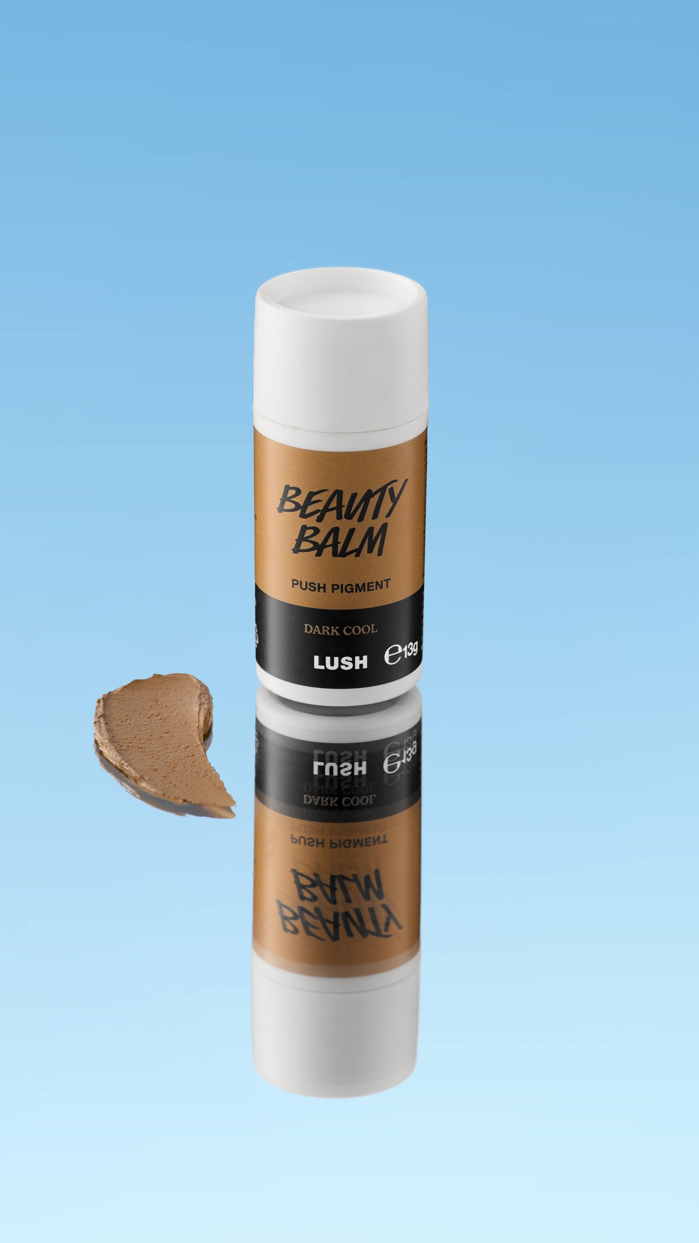 An angled product shot of a white and mustard-brown LUSH Beauty Balm Push Pigment stick in the shade Dark Cool. A thick swatch of the cool brown product rests next to the reflected stick, all against a bright blue background.