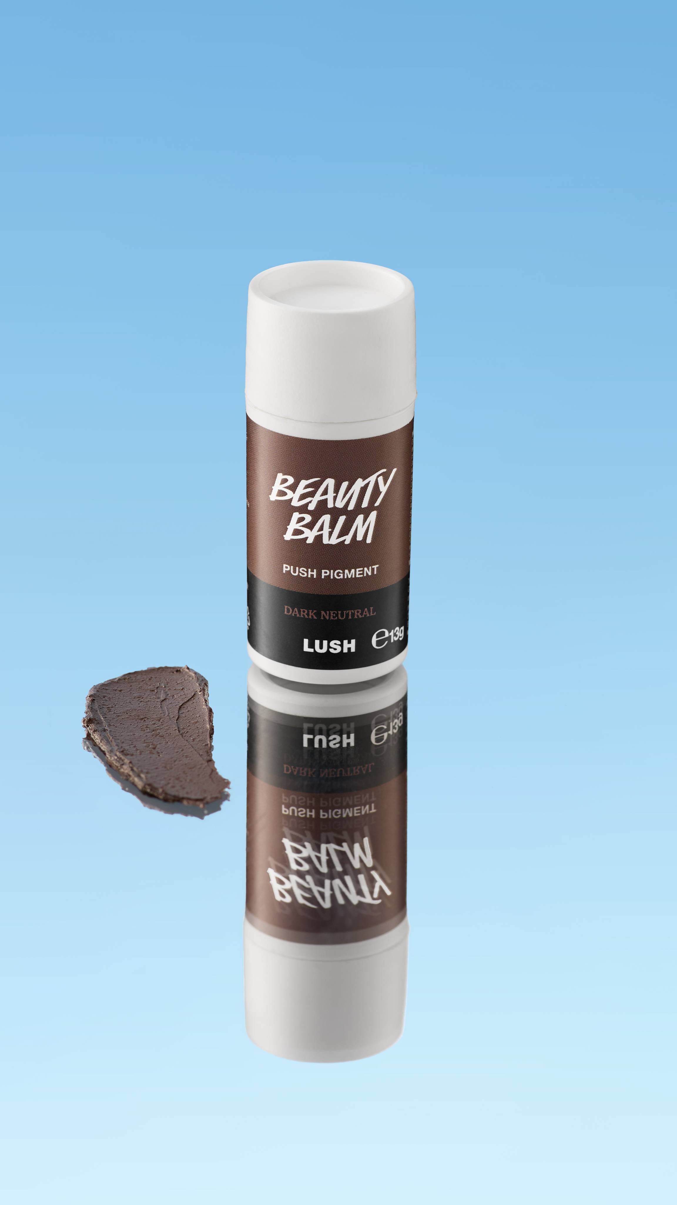 An angled product shot of a white and dark brown LUSH Beauty Balm Push Pigment stick in the shade Dark Neutral. A thick swatch of the deep brown product rests next to the reflected stick, all against a bright blue background.