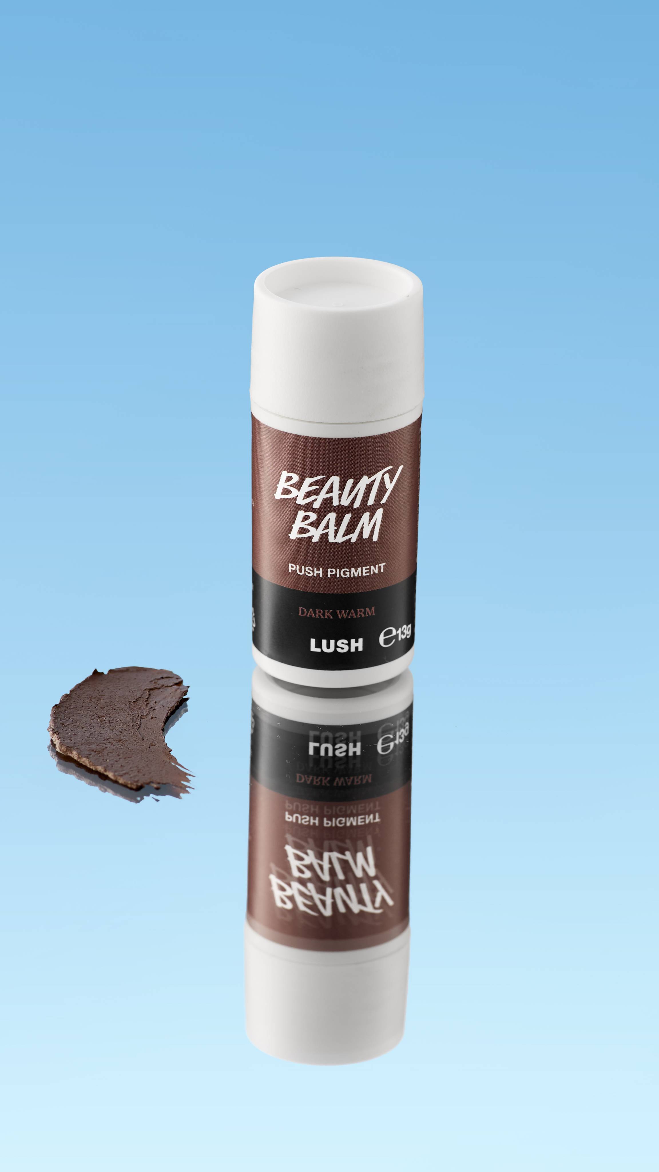 An angled product shot of a white and rich brown LUSH Beauty Balm Push Pigment stick in the shade Dark Warm. A thick swatch of the deep brown product rests next to the reflected stick, all against a bright blue background.