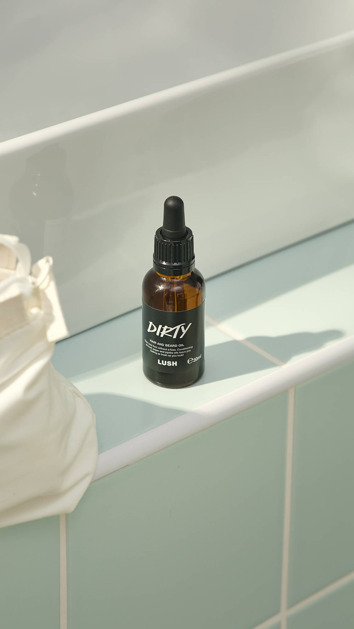 A small, amber glass bottle of "Dirty" hair and beard oil by Lush sits on a pale green tiled ledge. The bottle features a black label and a black pipette dropper lid. A white washbag is partially visible on the left.