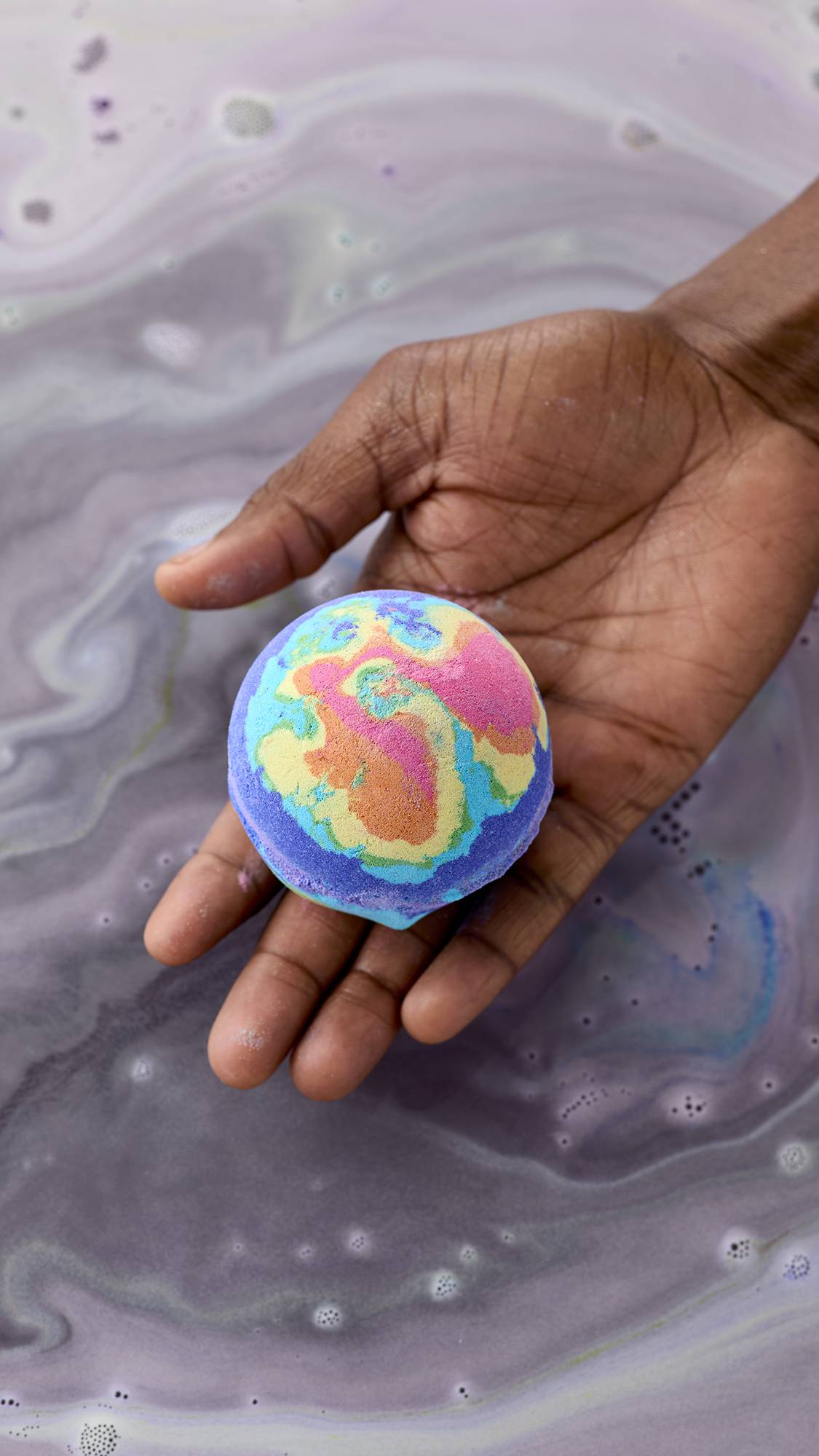 A model's hand holds a vibrant, multi-coloured "Diversity" bath bomb over swirling purple and grey bathwater. The bath bomb features a marbled pattern of pink, yellow, orange, green, and blue, resembling a miniature globe.