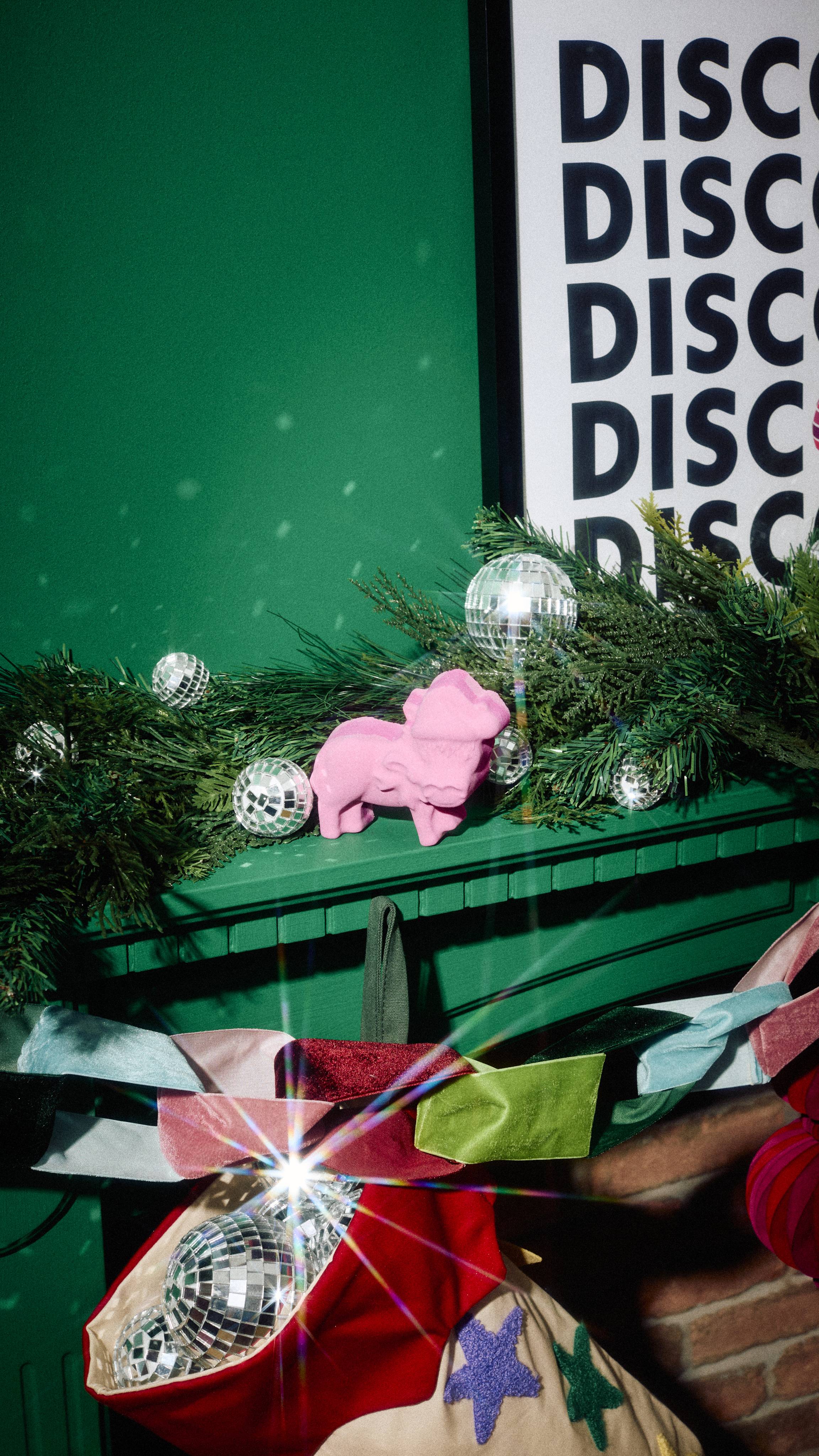 A pink donkey-shaped bath bomb sits on a green mantelpiece among pine garland and mini disco balls, above a colourful velvet bow garland and stocking filled with disco ball ornaments.