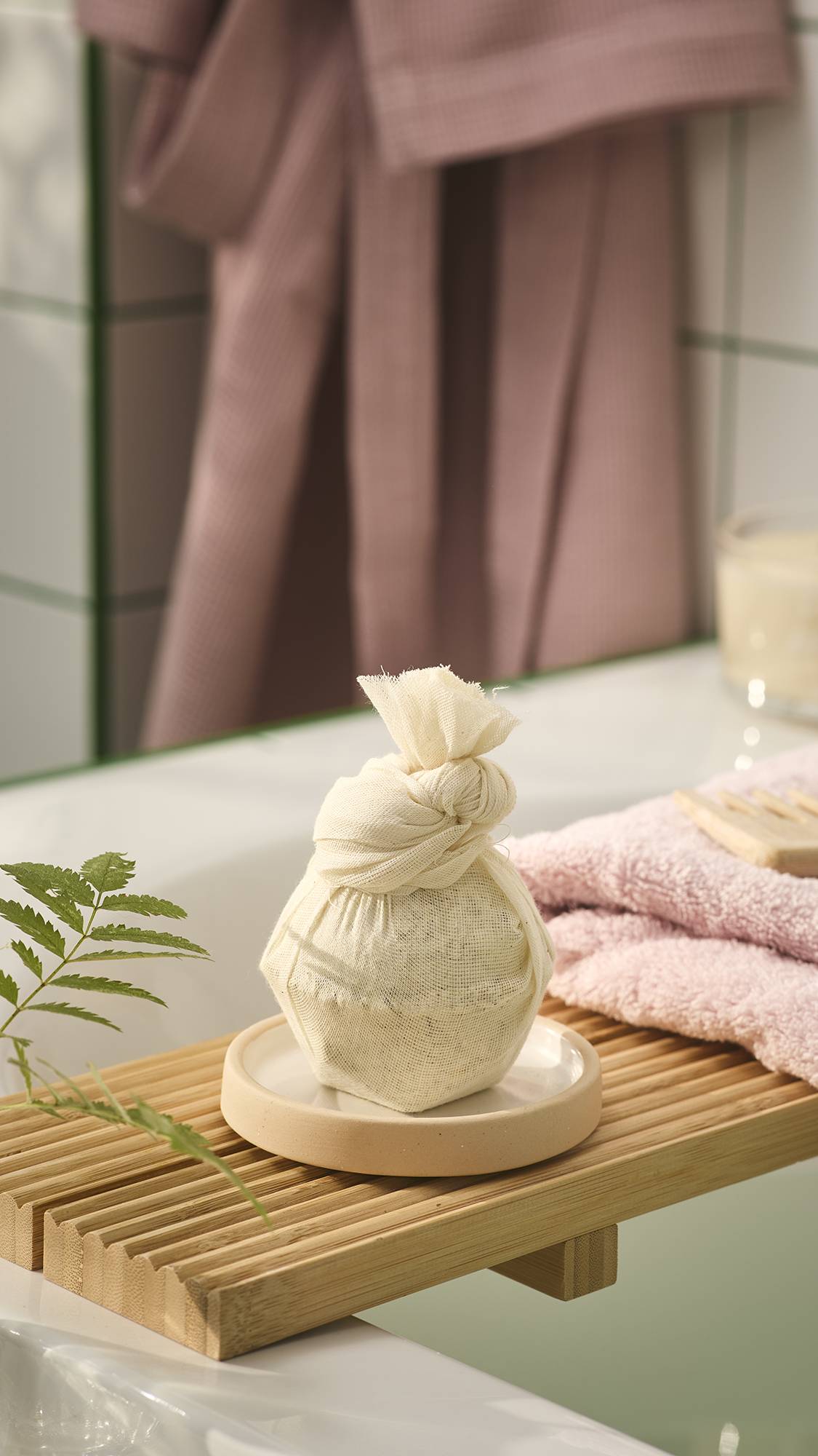 A bath bomb wrapped in a cream muslin cloth pouch tied with a knot sits on a small dish atop a wooden bath caddy, set against a bathroom background with a pink dressing gown.