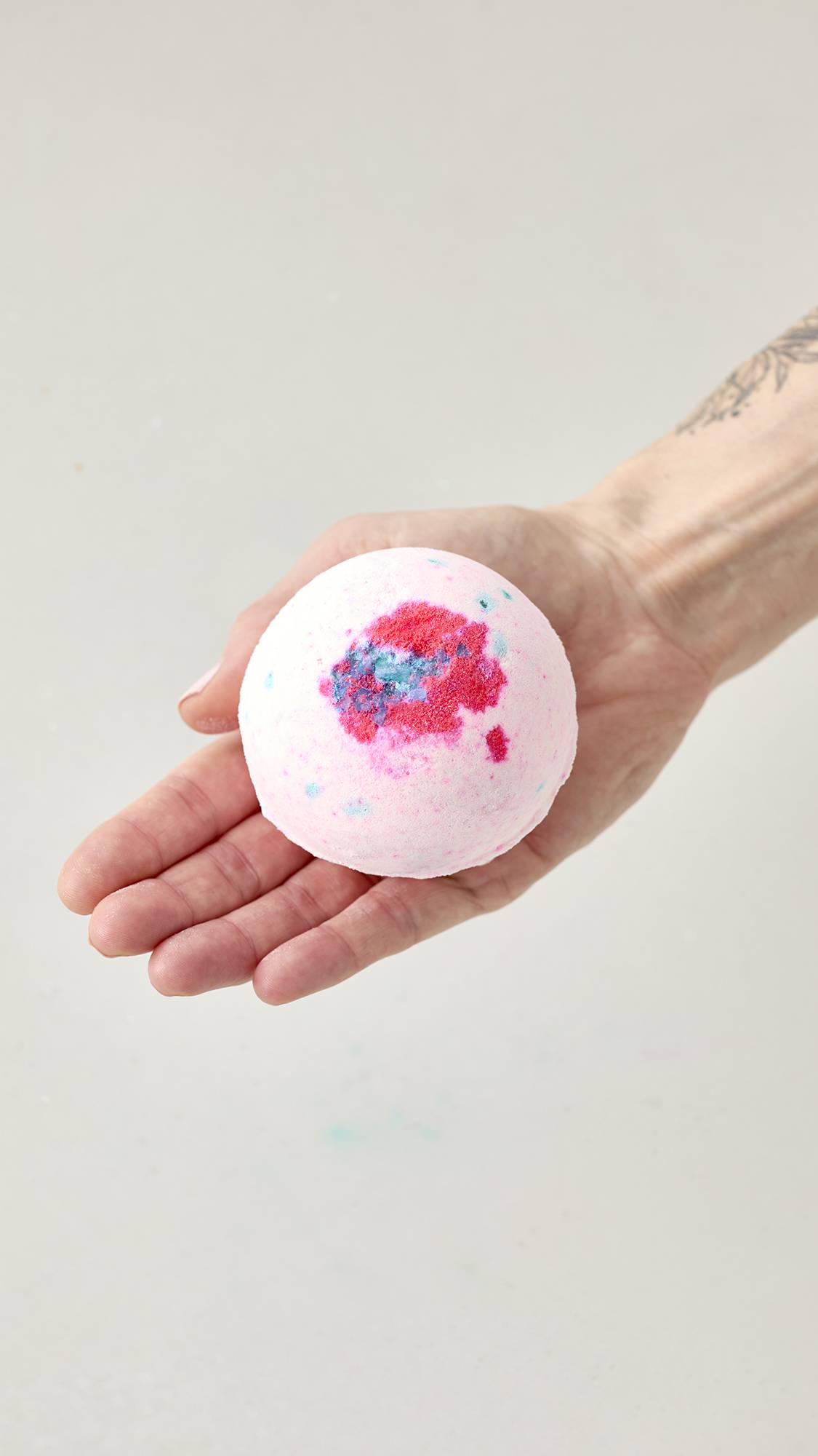 A hand holds the dry, faceted Equity bath bomb. The geometric product features crisp, color-blocked sections of pastel pink, bright blue, and white against a plain background.