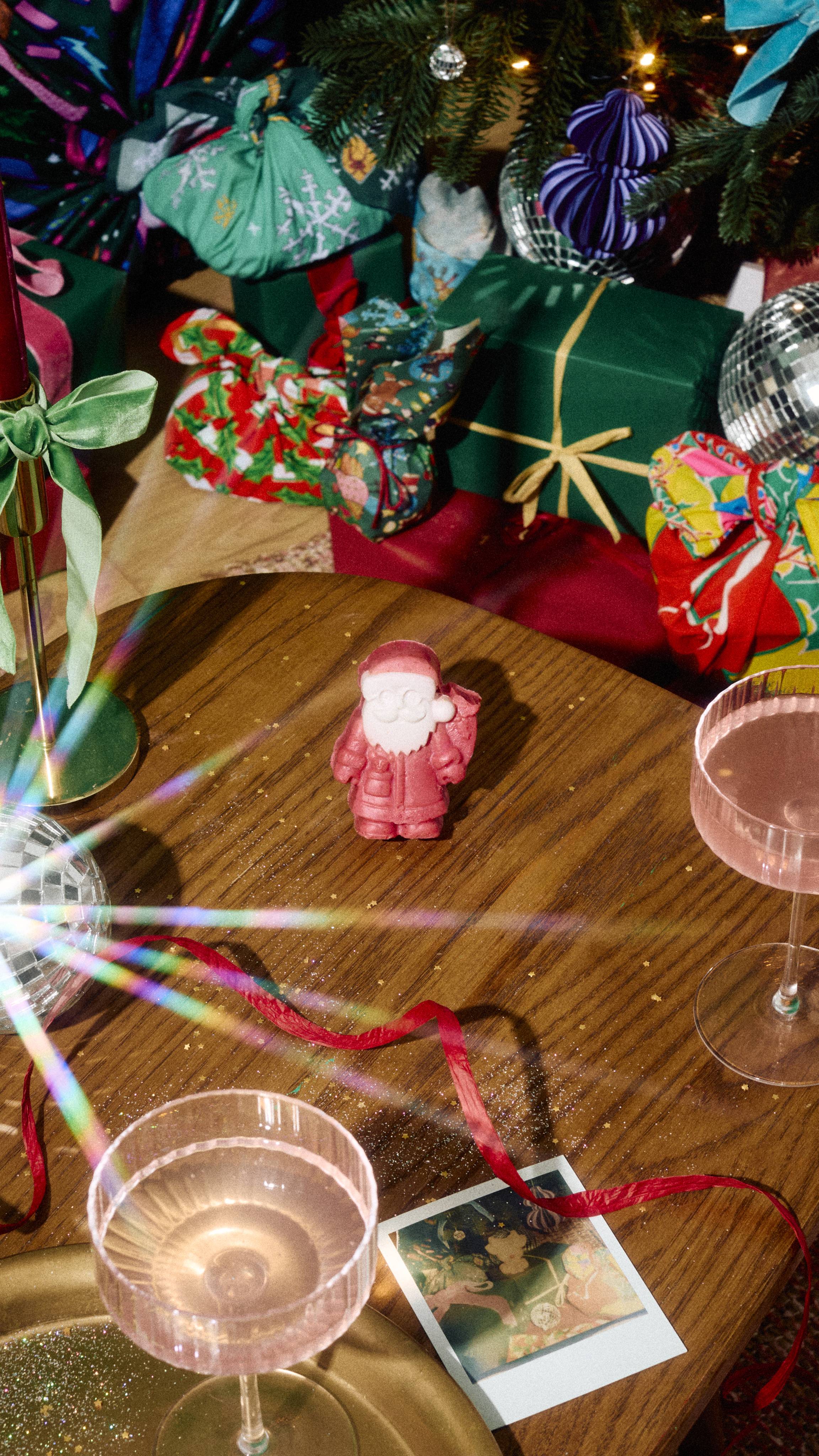 A moulded Santa Claus soap bar stands upright on a wooden table, surrounded by glasses of pink drink, red ribbon, and festive gifts under a decorated Christmas tree.