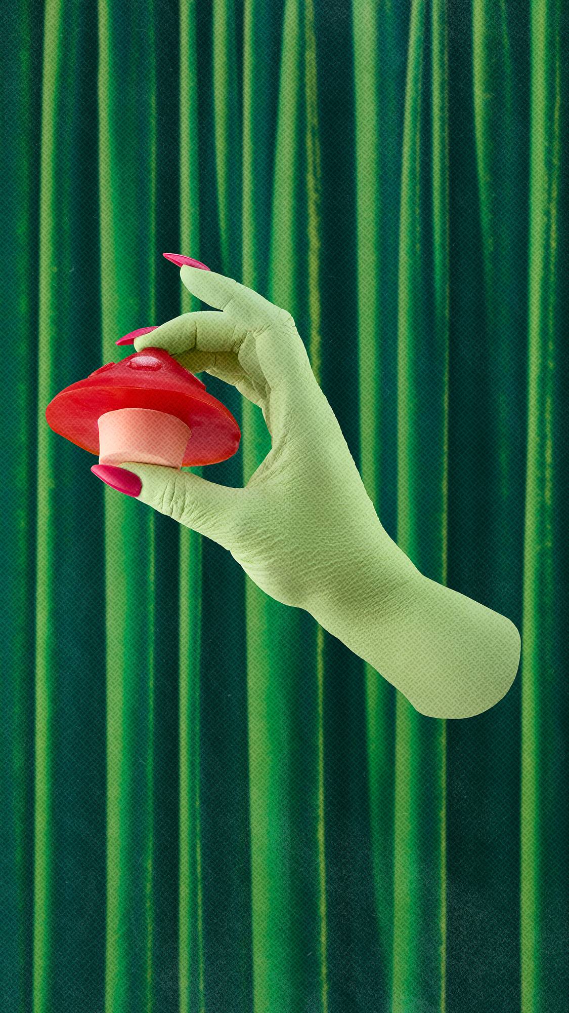 A green-skinned hand with long pink nails holds a toadstool-shaped soap with a red cap and cream stem, set against dark green velvet curtains.