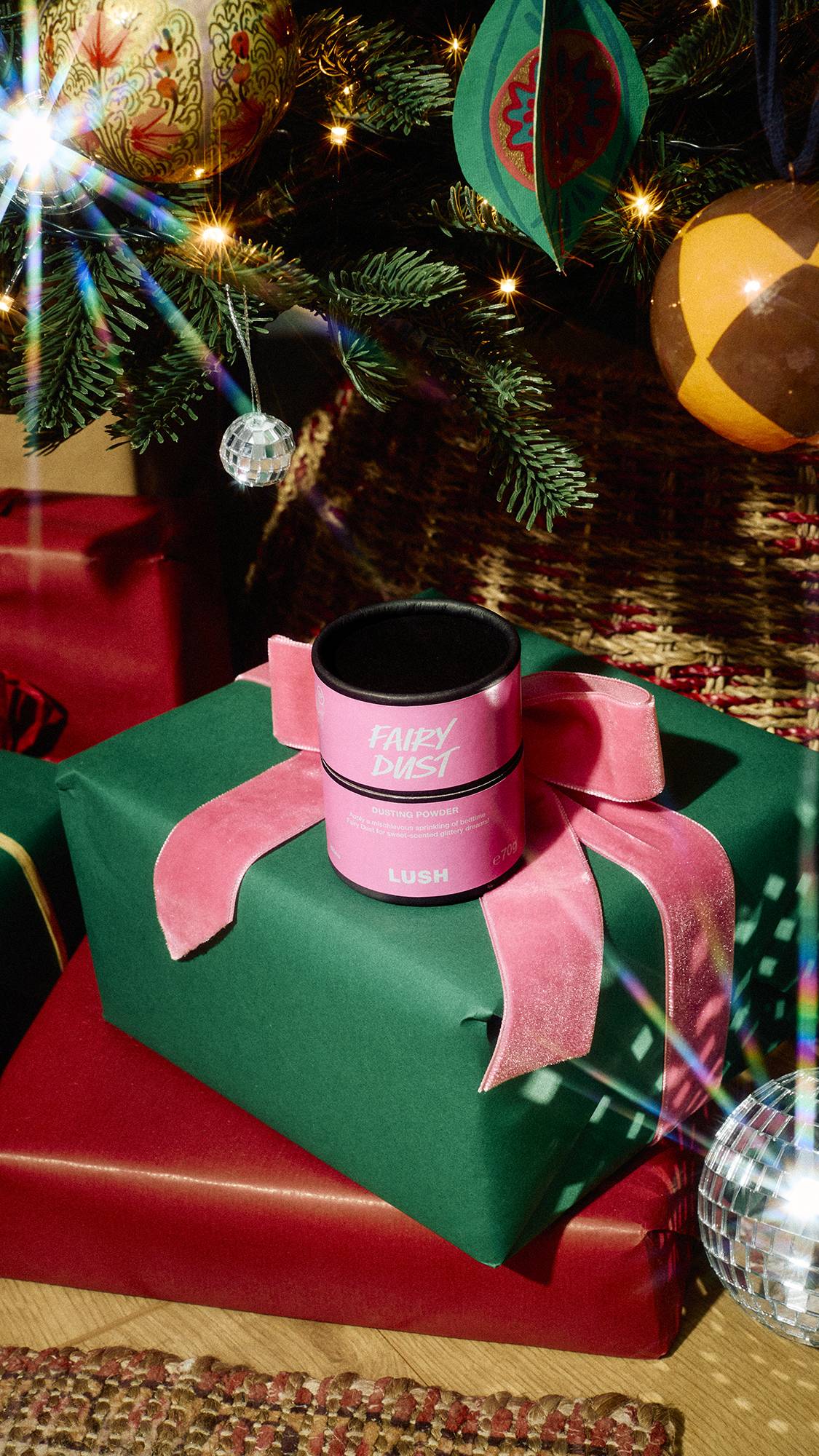 A pink and black cardboard tub of Lush Fairy Dust dusting powder rests on a green gift tied with a pink velvet ribbon beneath a decorated Christmas tree.